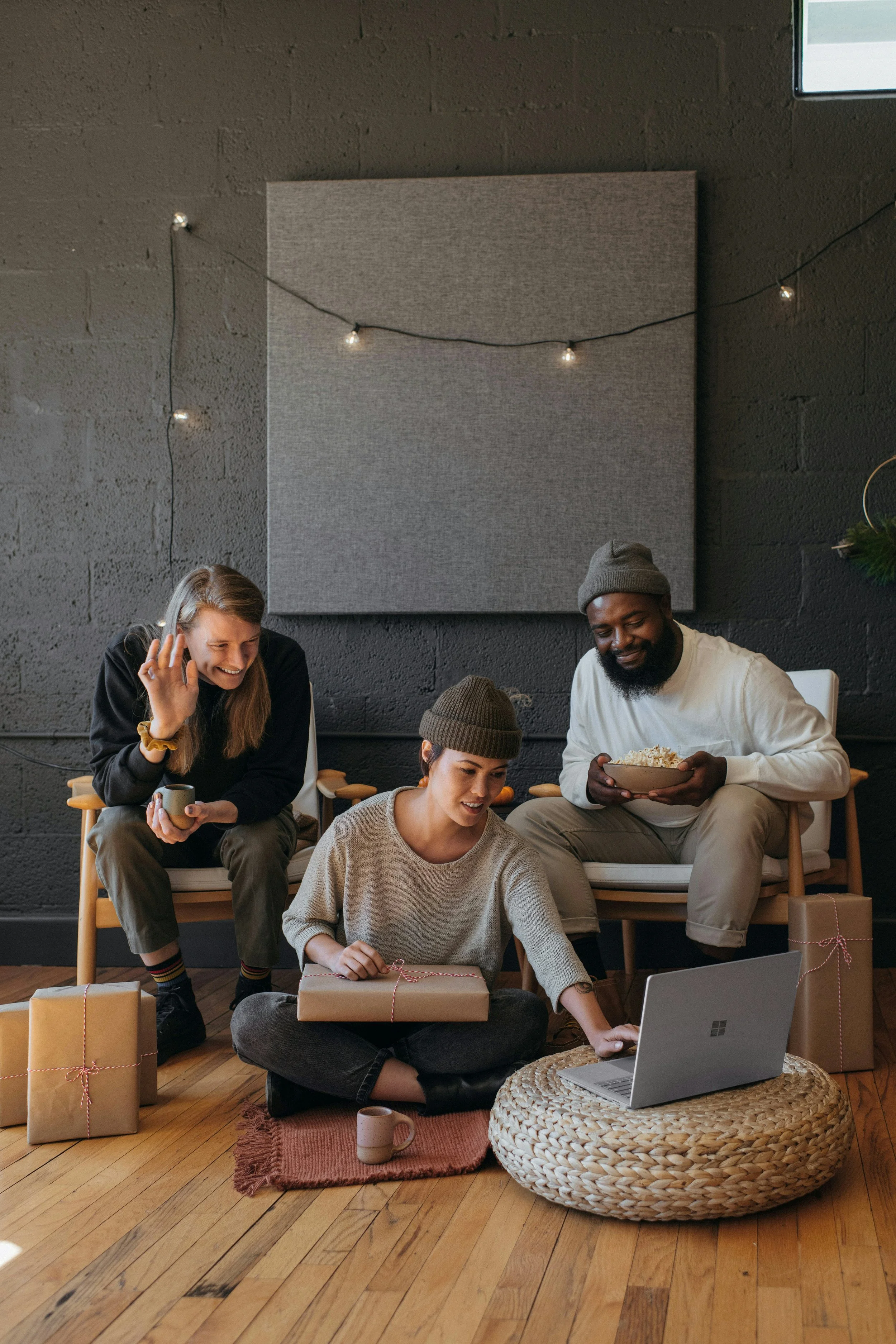 Group of four friends celebrating Christmas indoors, with gifts, laptops, and warm clothing, sitting on the floor and on a ledge against a grey wall with string lights.