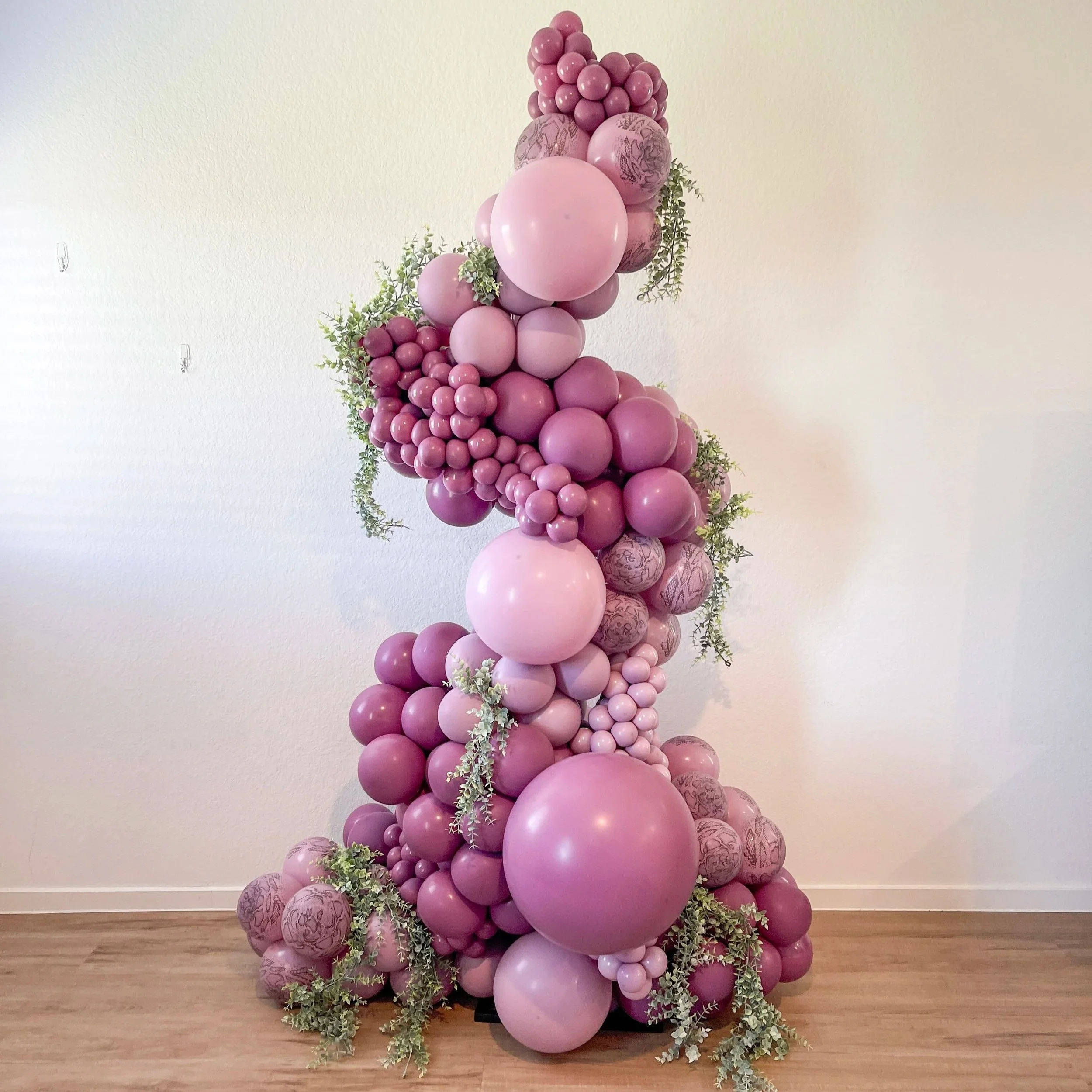 A large balloon arrangement with pink and patterned balloons, accented with greenery, set against a plain white wall and wooden floor.