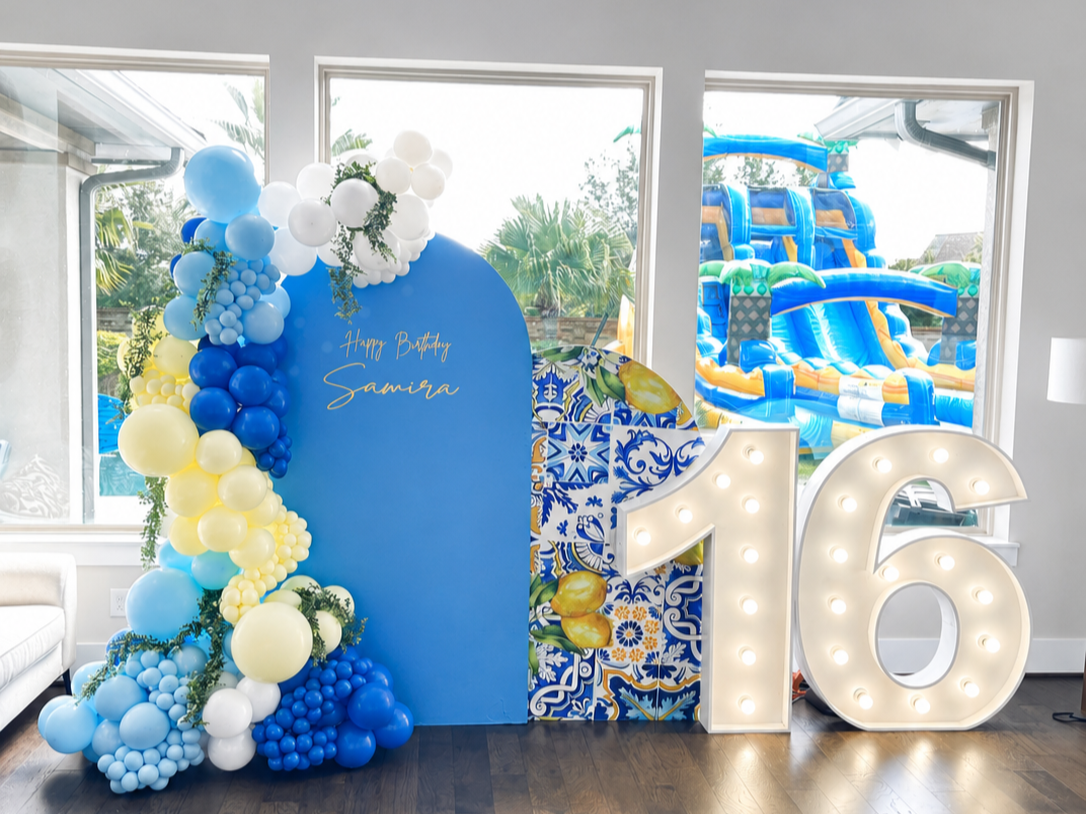 Decorative birthday celebration with balloons in blue, yellow, and white, a large illuminated number 16, and a blue sign that says 'Happy Birthday Samira.' Large inflatable water slides are visible outside through the windows.