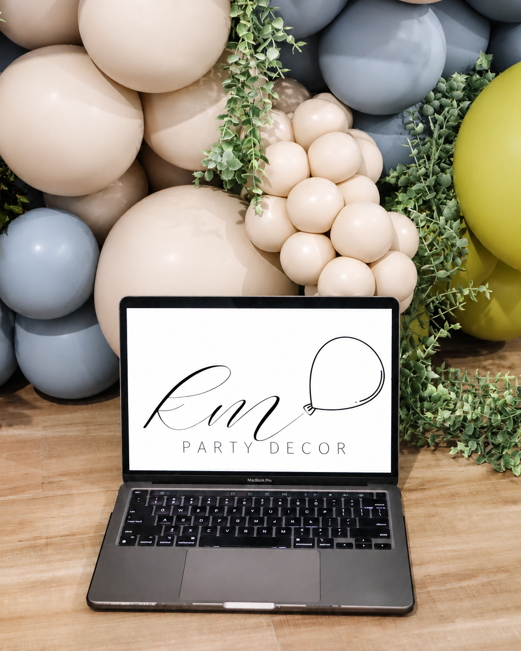 Laptop on wooden table with a digital screen displaying the logo for 'km Party Decor,' surrounded by pastel-colored balloons and green foliage.