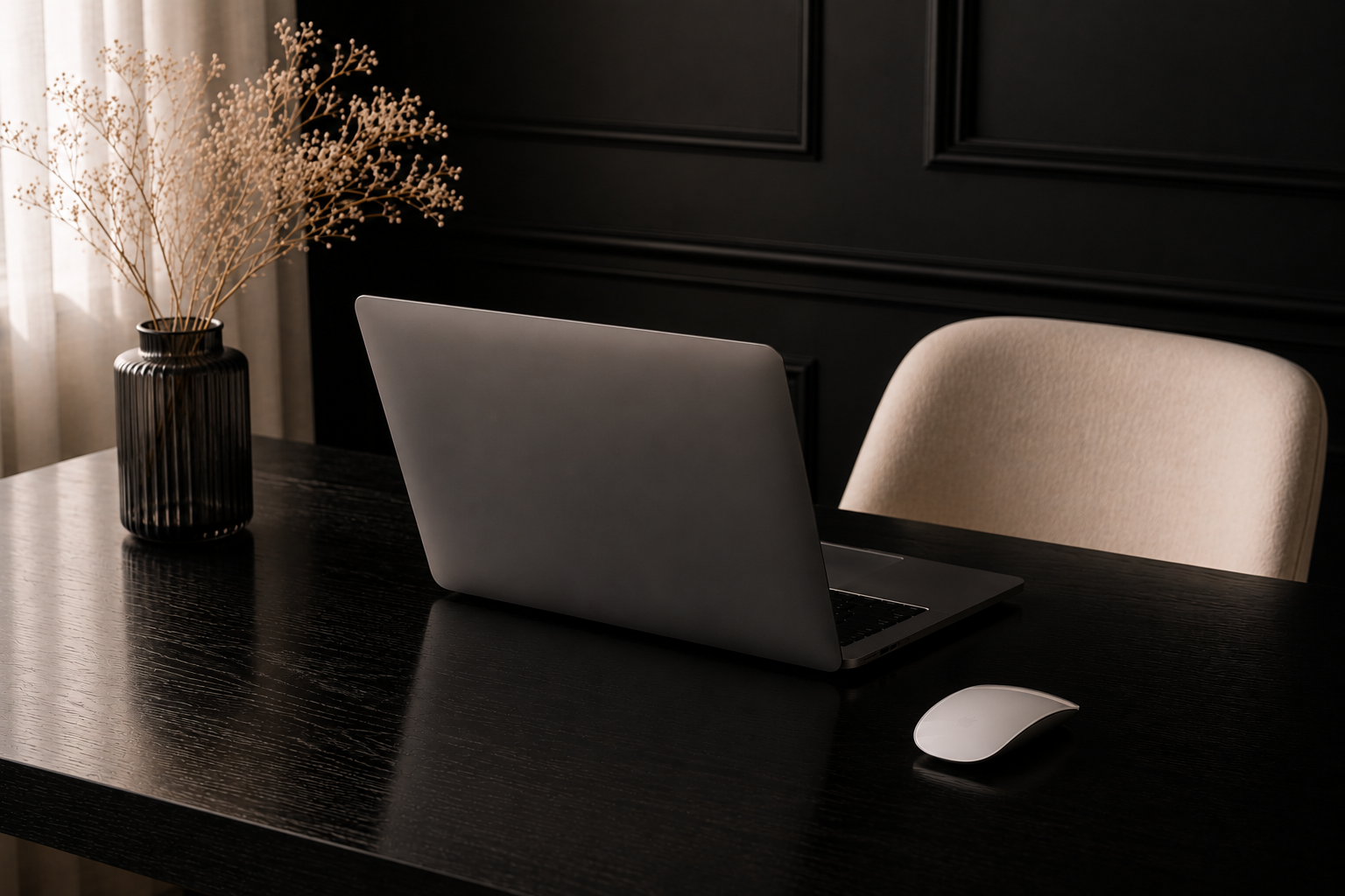 A dark wooden desk with a silver laptop, a white computer mouse, a black vase with dried flowers, a beige chair, and black wall paneling in the background.