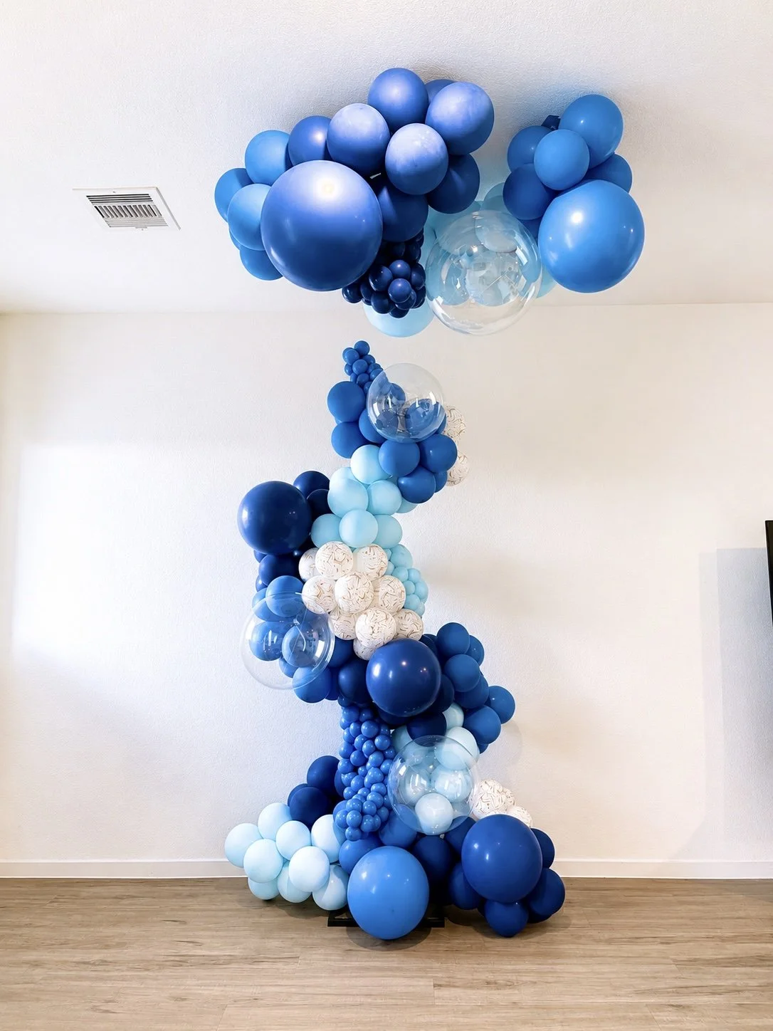 Blue balloon arrangement with varying shades, clear and white balloons, forming a large decorative display in a room with light-colored walls and wooden flooring.