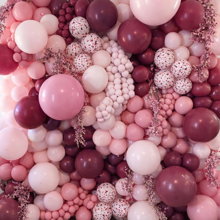 A colorful balloon arrangement with shades of pink, white, and burgundy, featuring various sizes and some with confetti patterns, accented with small pink flowers.