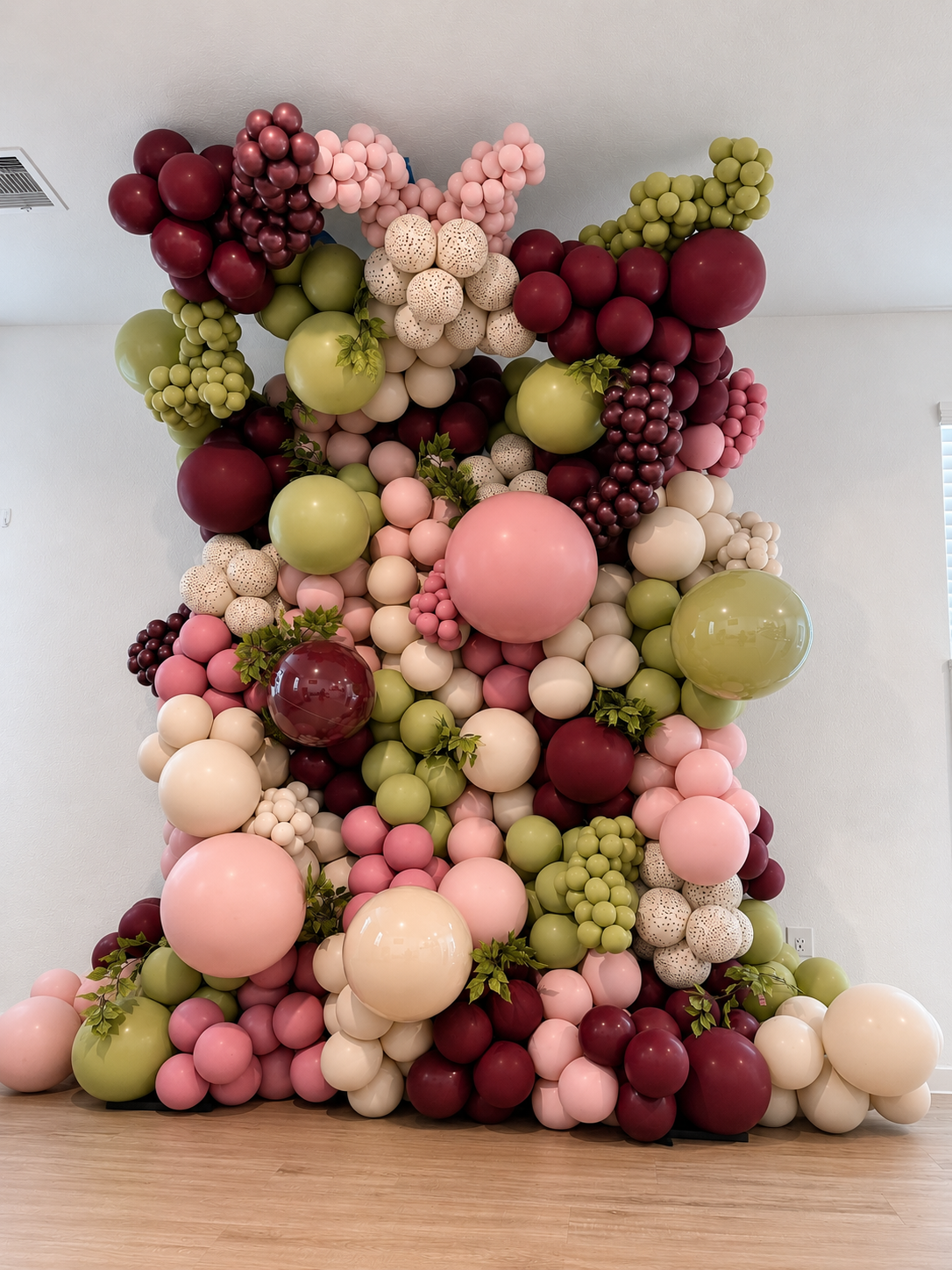 A large decorative balloon arrangement consisting of pink, green, white, burgundy, and beige balloons in various sizes, with some green leaves interspersed, set against a plain white wall.
