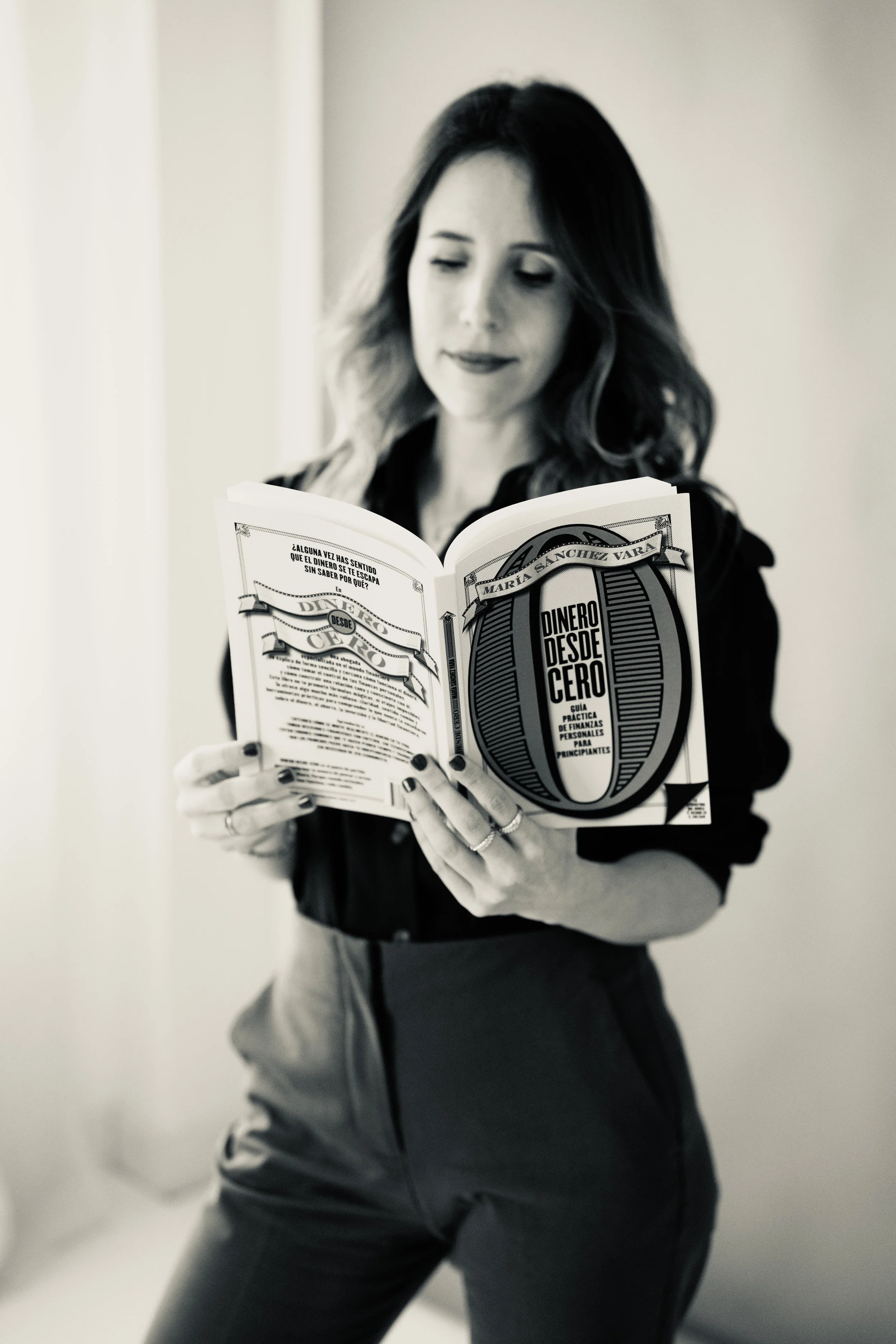 Mujer leyendo un libro titulado 'Dinero desde cero' mientras está de pie en un espacio interior.