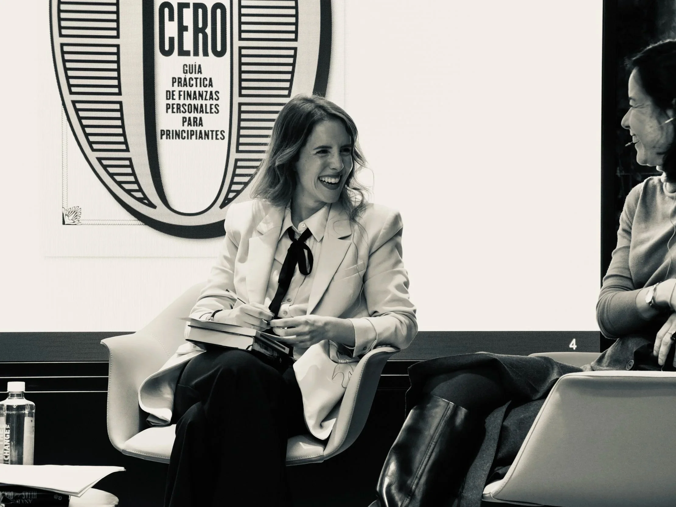 Dos mujeres conversando en una sala de reuniones, una de ellas sostiene una libreta y un bolígrafo. Detrás de ellas hay un cartel que dice 'CERO GUÍA PRÁCTICA DE FINANZAS PERSONALES PARA PRINCIPIANTES'.