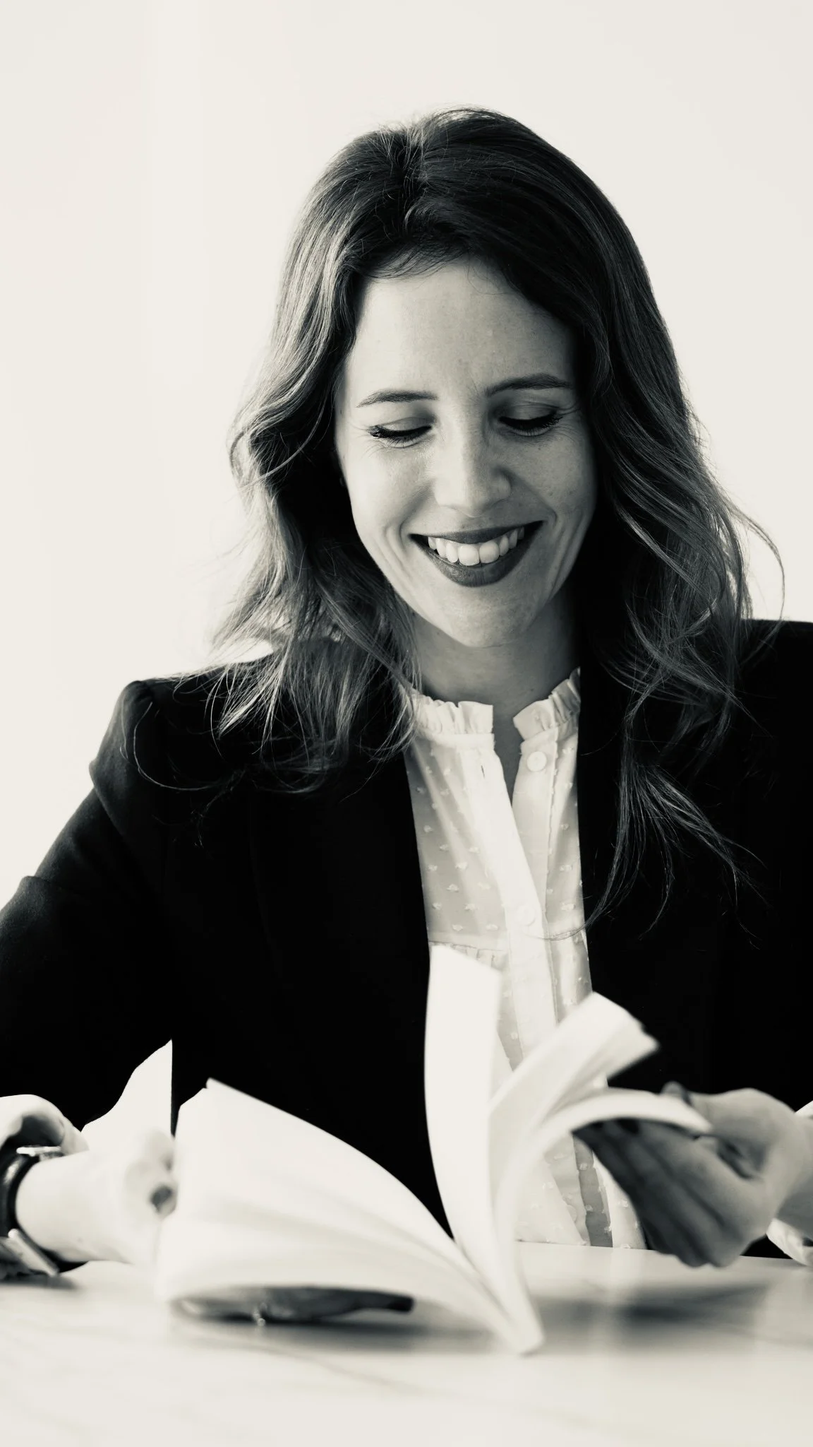 Mujer sonriendo mientras lee un libro en una mesa. La imagen es en blanco y negro.