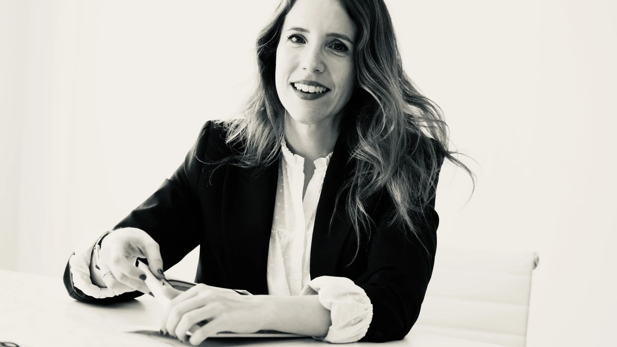 Mujer con cabello largo y rizado, vestida con chaqueta oscura y blusa blanca, sonriendo y escribiendo en un cuaderno en una mesa blanca.