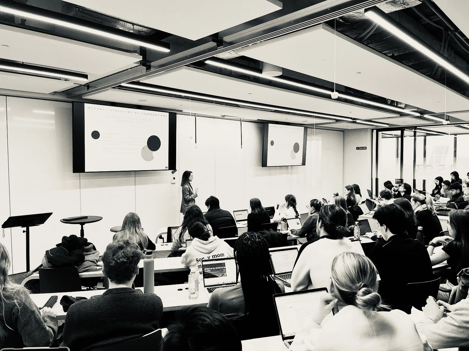 Una mujer dando una presentación a un grupo de personas en un aula de clases moderna con múltiples pantallas y estudiantes usando laptops.