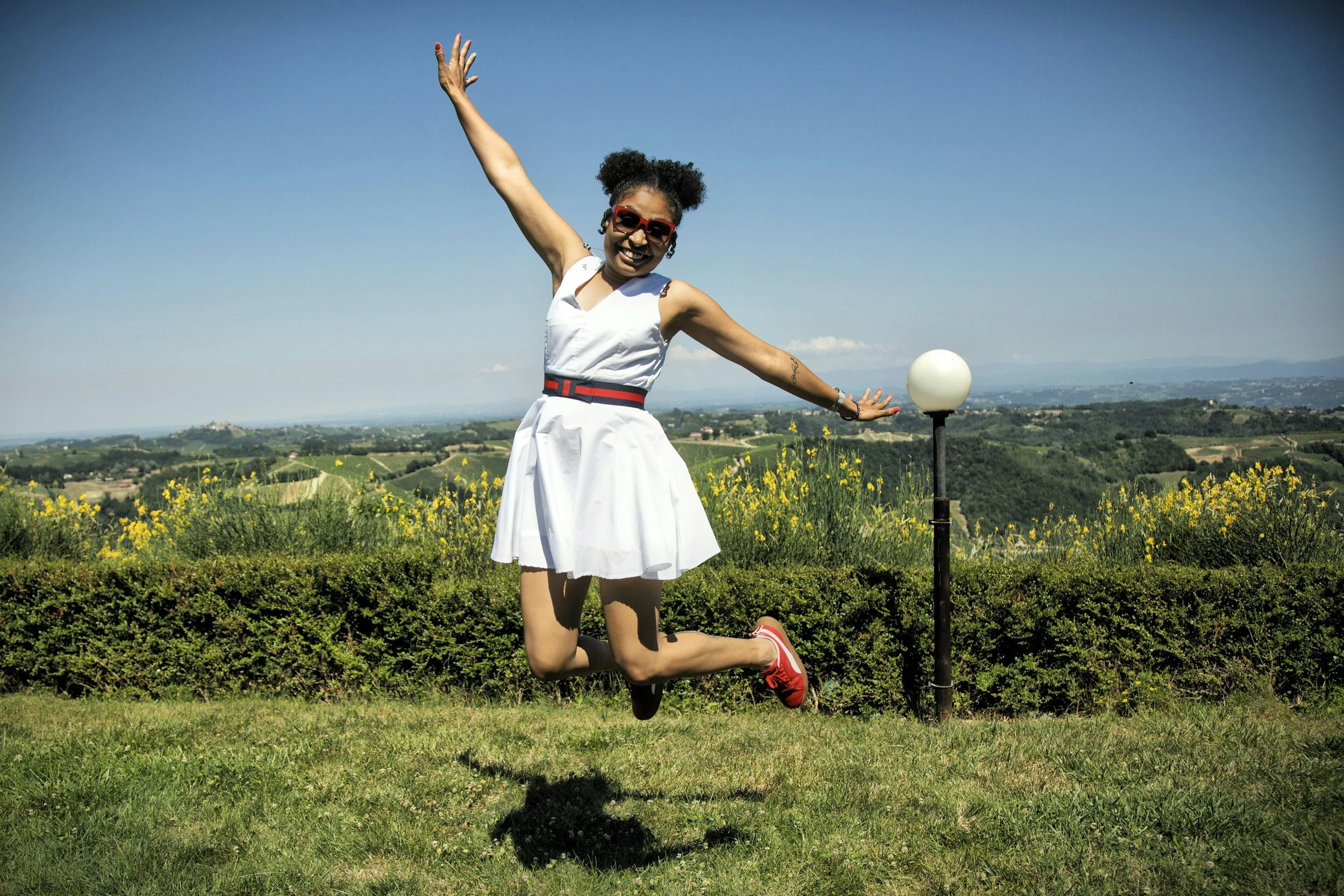 A woman with curly hair, sunglasses, red sneakers, and a white dress with a red and black belt, is outdoors on a grassy hill, kneeling on one knee, with one arm raised and the other arm extended to the side. A lamp post and yellow flowers are in the foreground, with rolling hills and blue sky in the background.