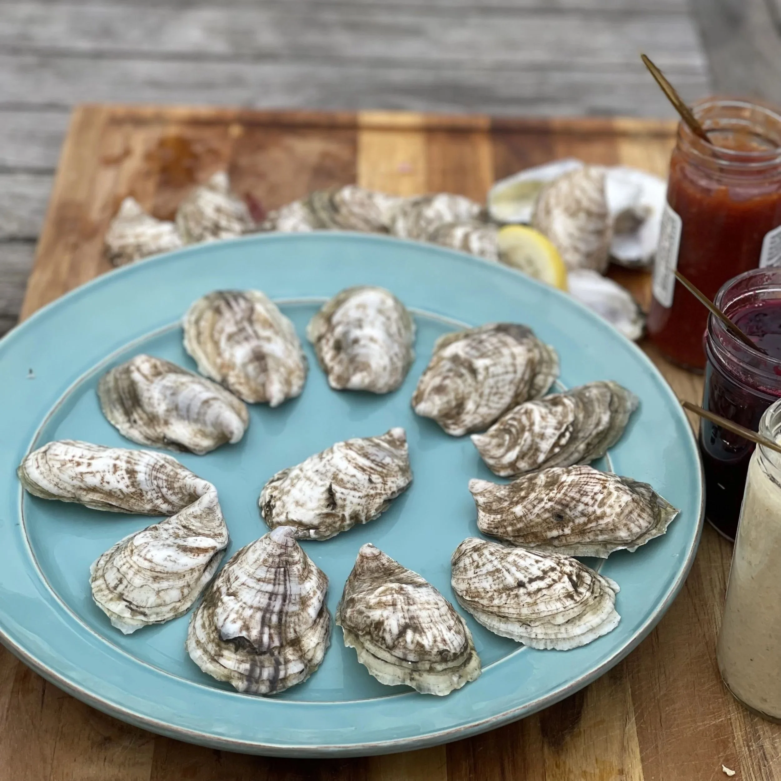 A blue plate filled with cooked oysters on a wooden table, with a wooden tray and jars of sauce in the background.