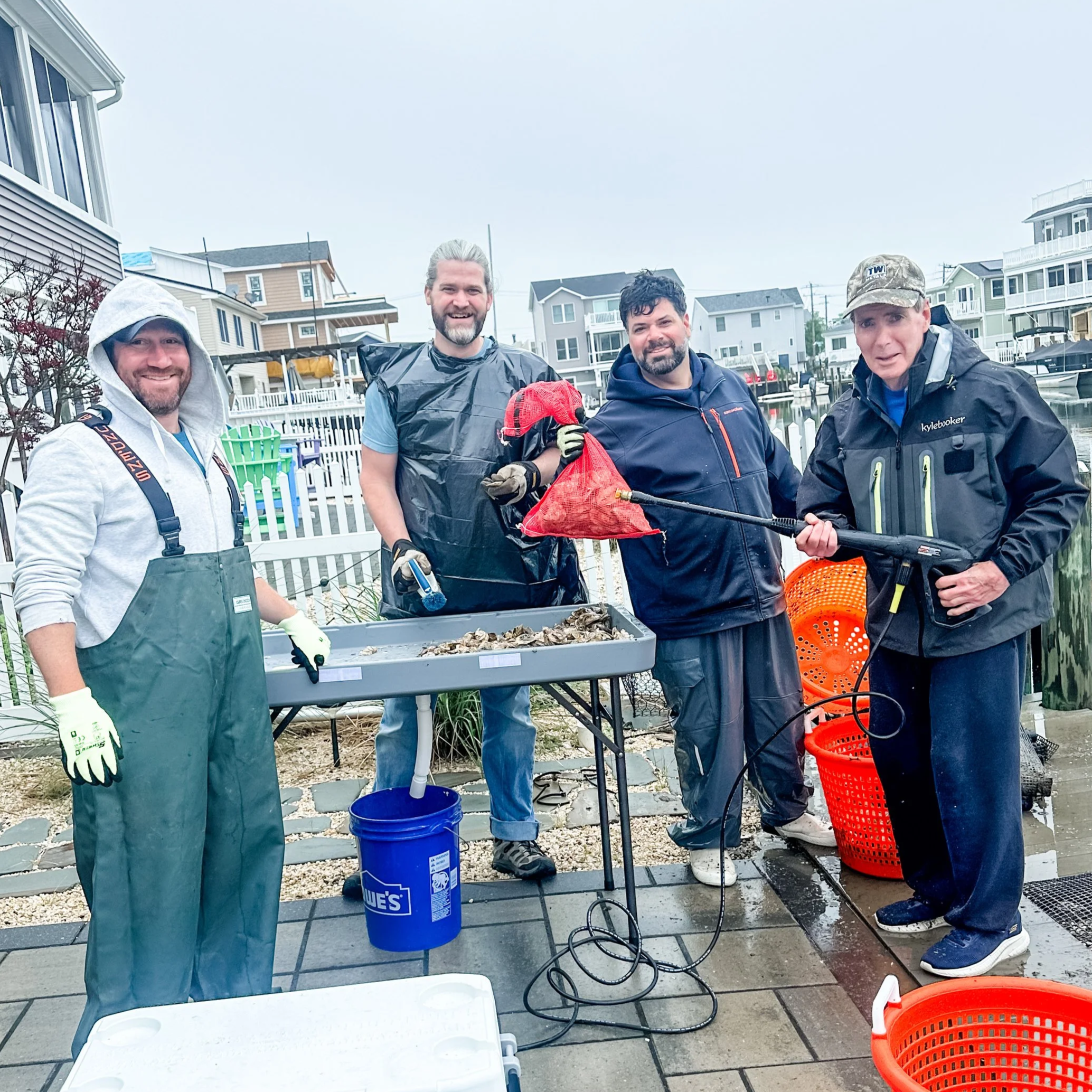 Four men standing outdoors near a body of water, holding harvest bags, dressed in outdoor gear including waterproof jackets, with houses in the background, rainy weather, and fishing equipment around.