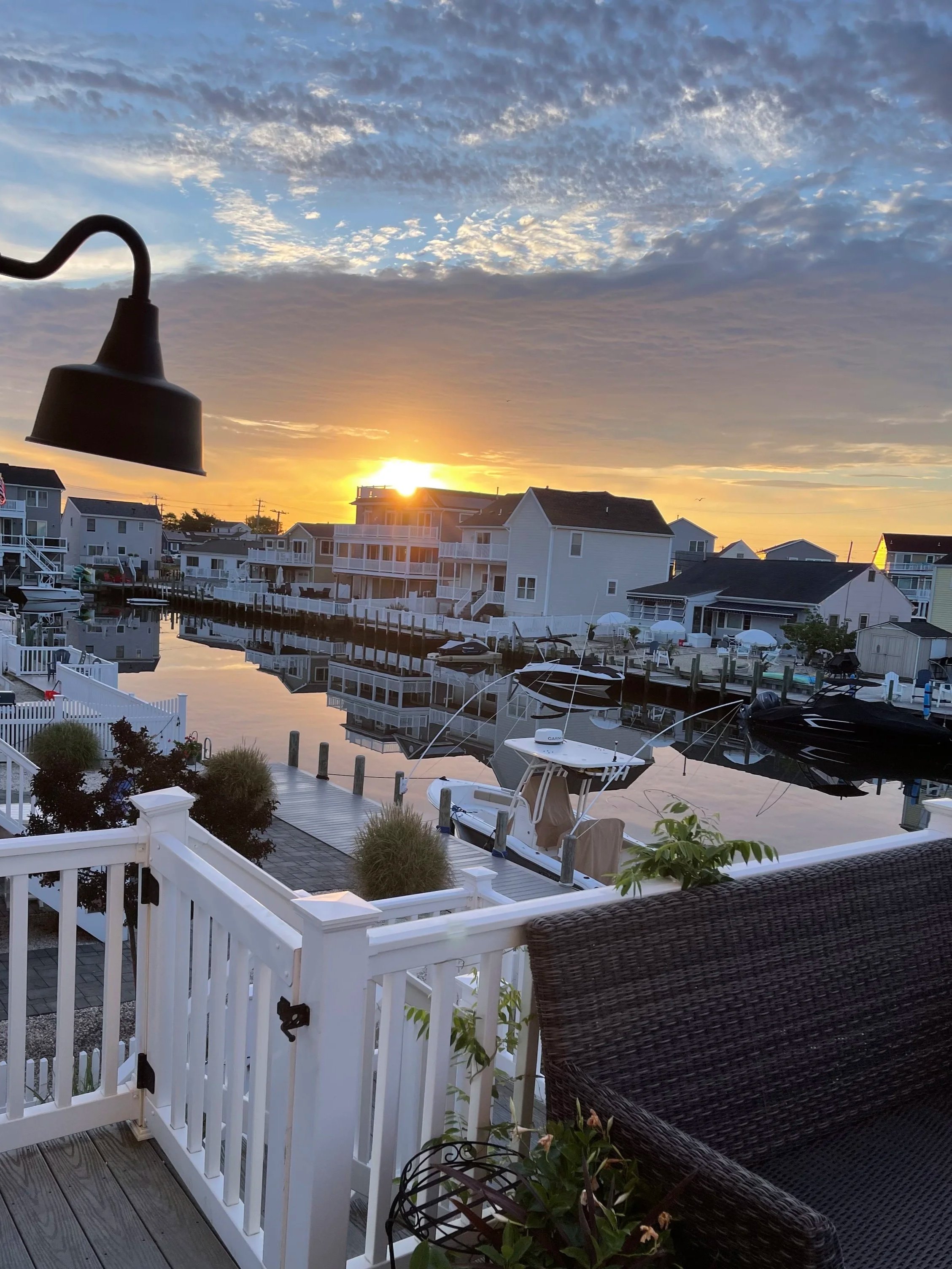View of a canal with boats, residential houses, and a dock at sunset, seen from a balcony with a black wicker chair and white railing.