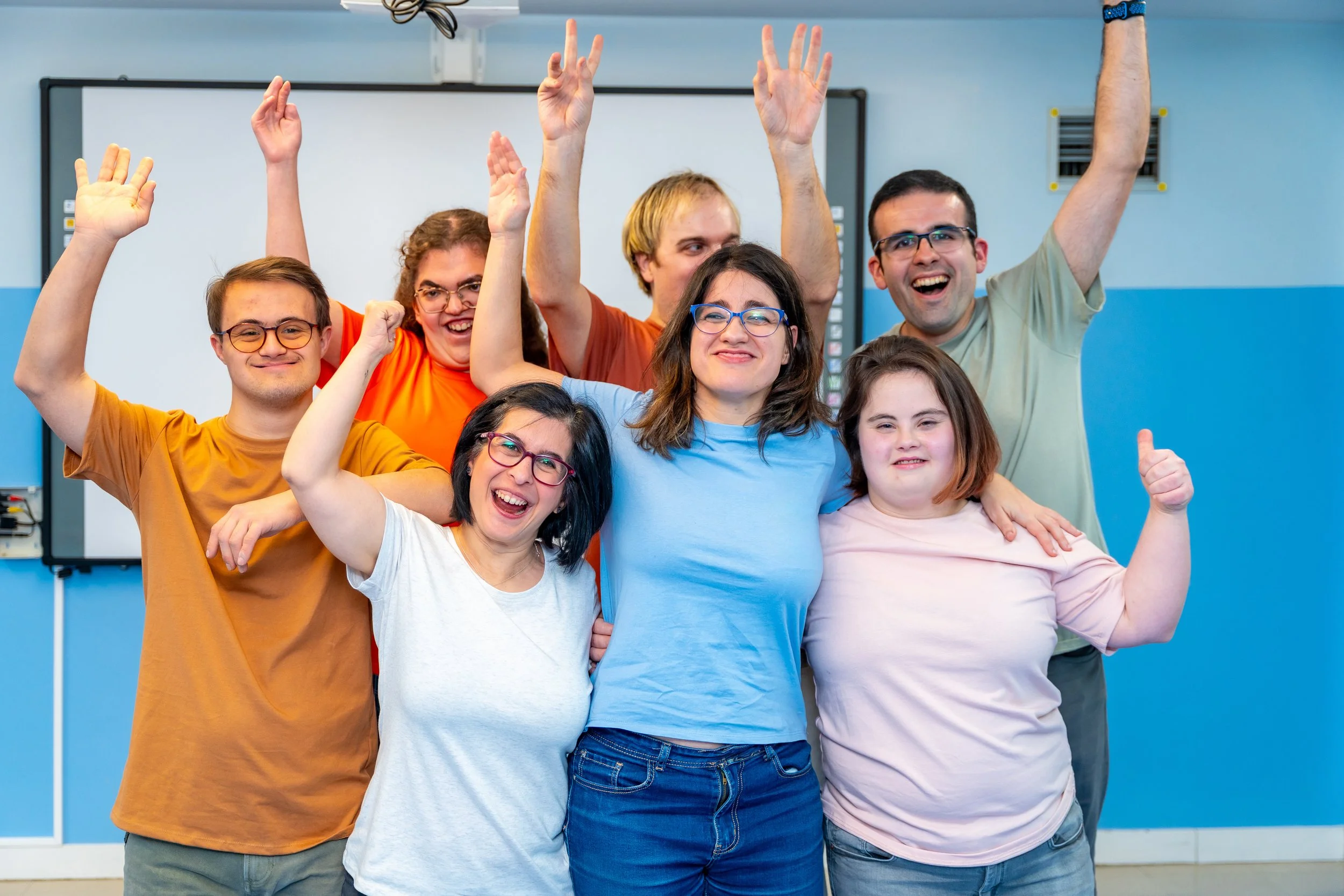 A group of seven diverse young adults celebrating together in a classroom, smiling, with some raising their hands and giving thumbs up.