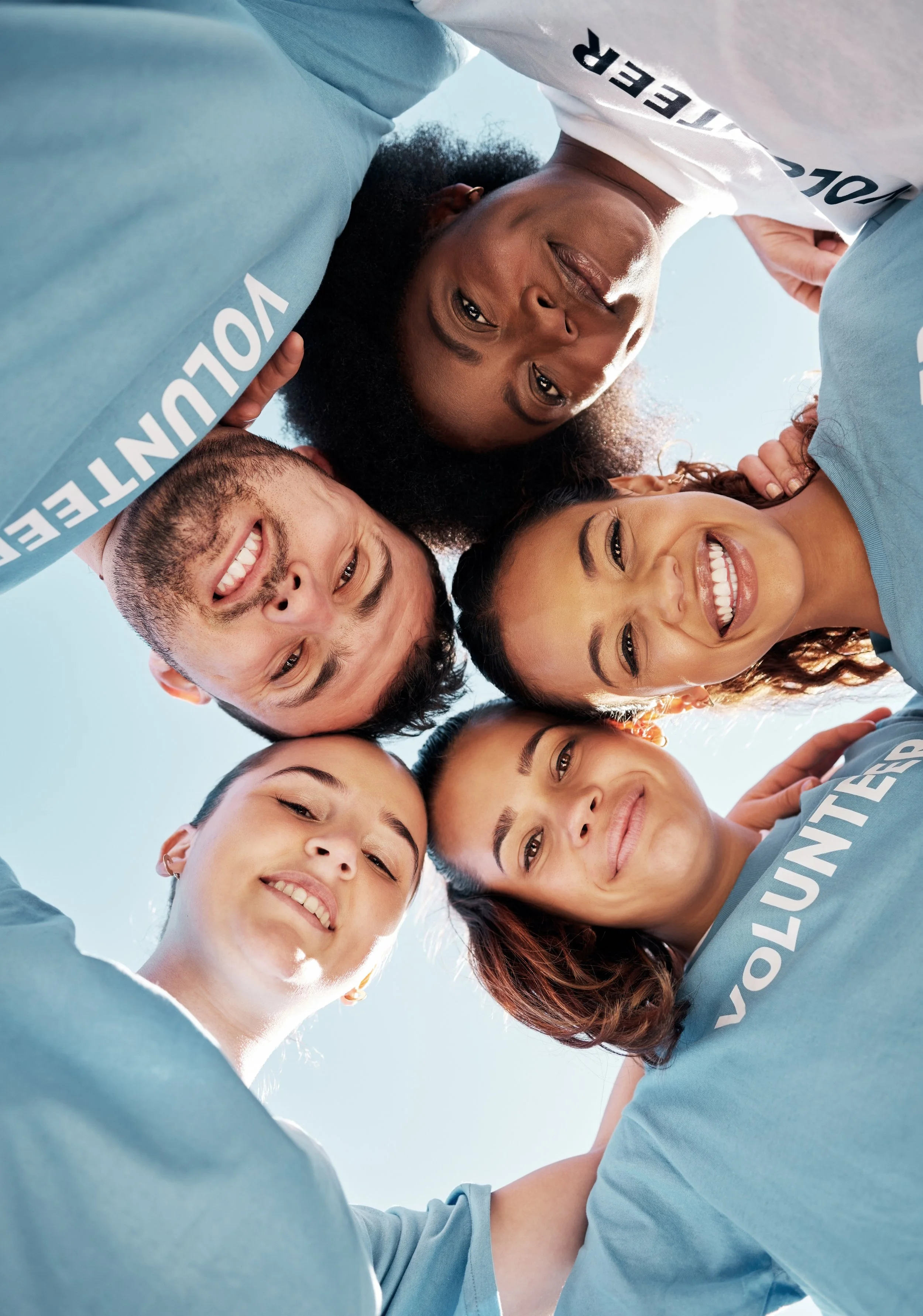 A diverse group of young adults in casual clothing forming a circle with their heads close together, smiling and looking down at the camera, with a clear blue sky background.