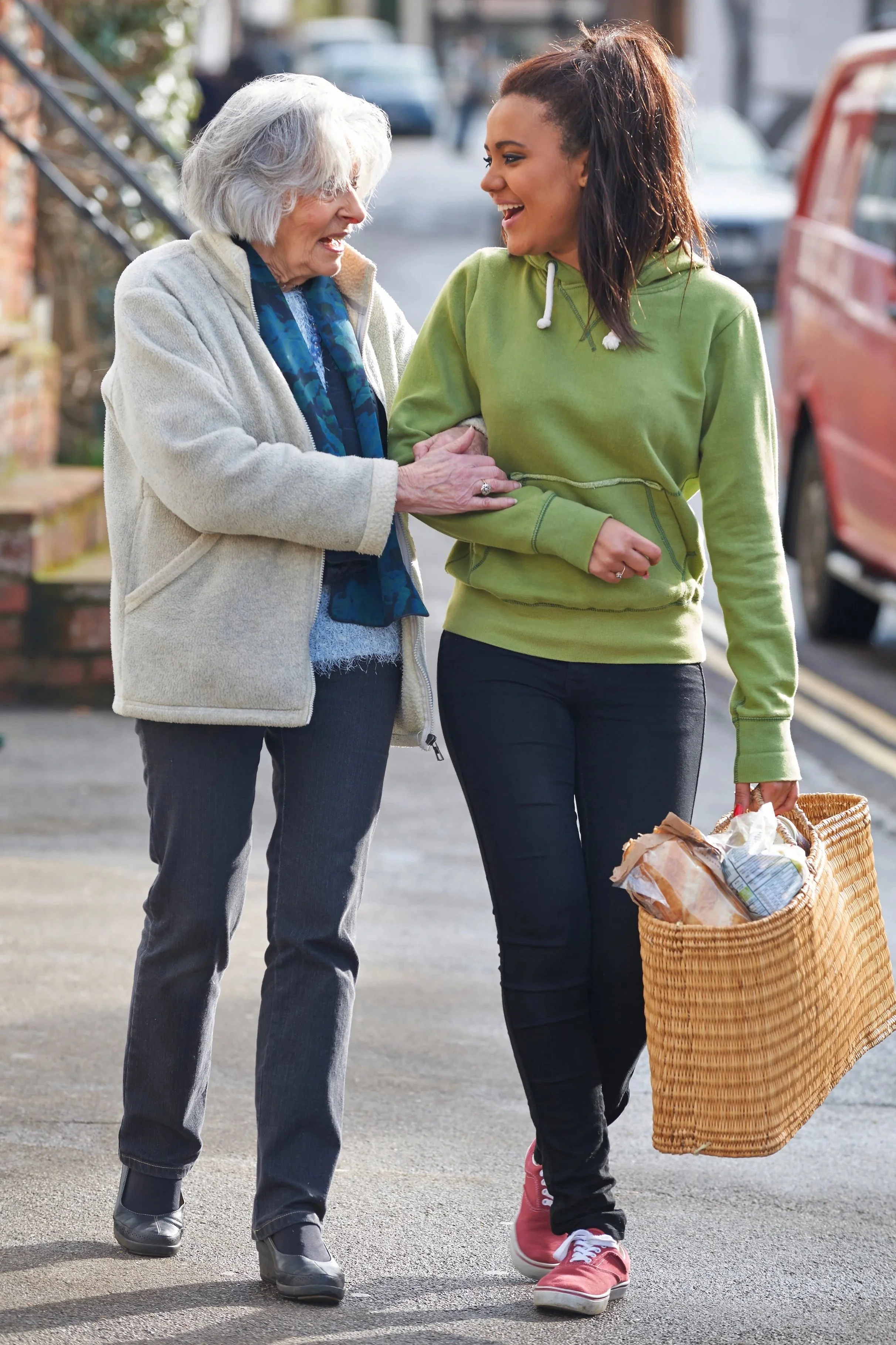 A young woman and an elderly woman are smiling and holding hands while walking on a city street. The young woman is carrying a wicker basket filled with groceries.