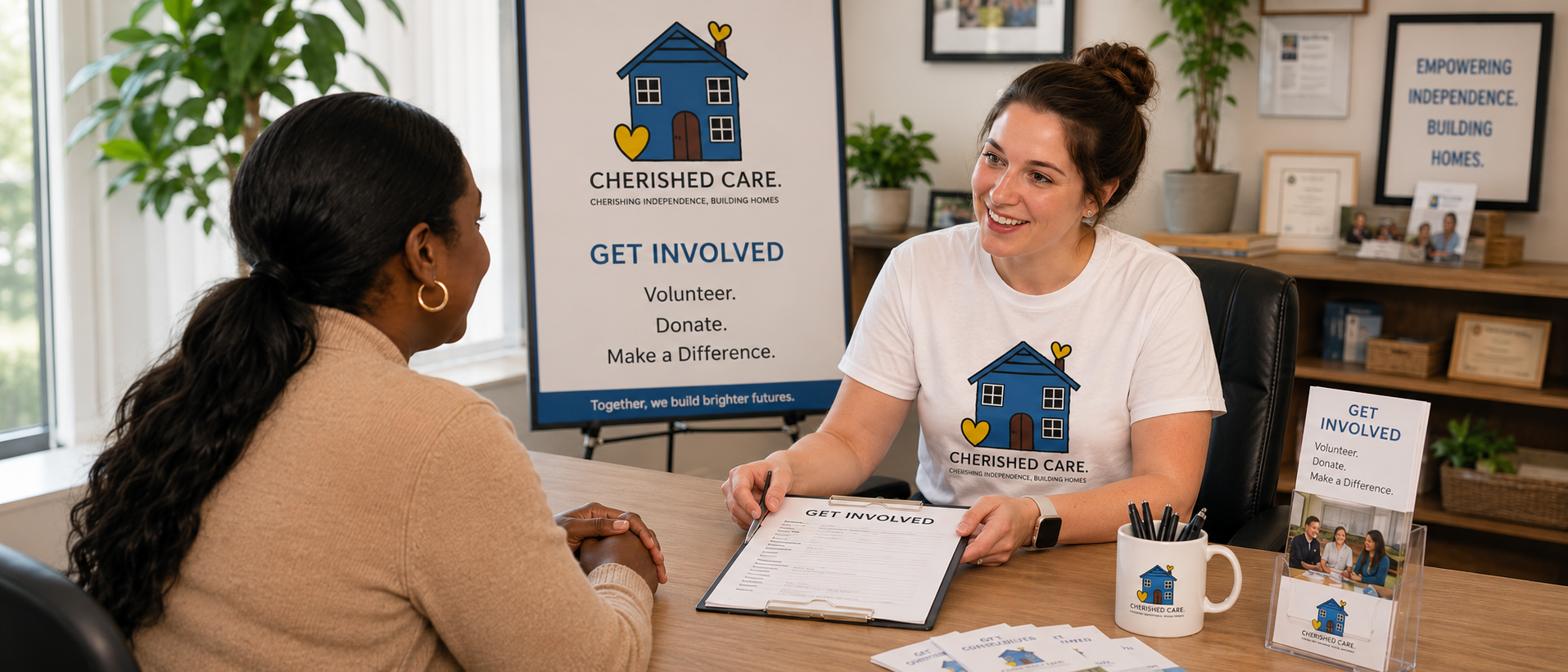 Two women engaged in a conversation at a table in an office setting, with promotional materials and a banner for Cherished Care in the background. The woman on the right, wearing a white t-shirt with the Cherished Care logo, is smiling and holding a clipboard labeled 'Get Involved.' The woman on the left, with dark hair in a ponytail, is listening attentively.