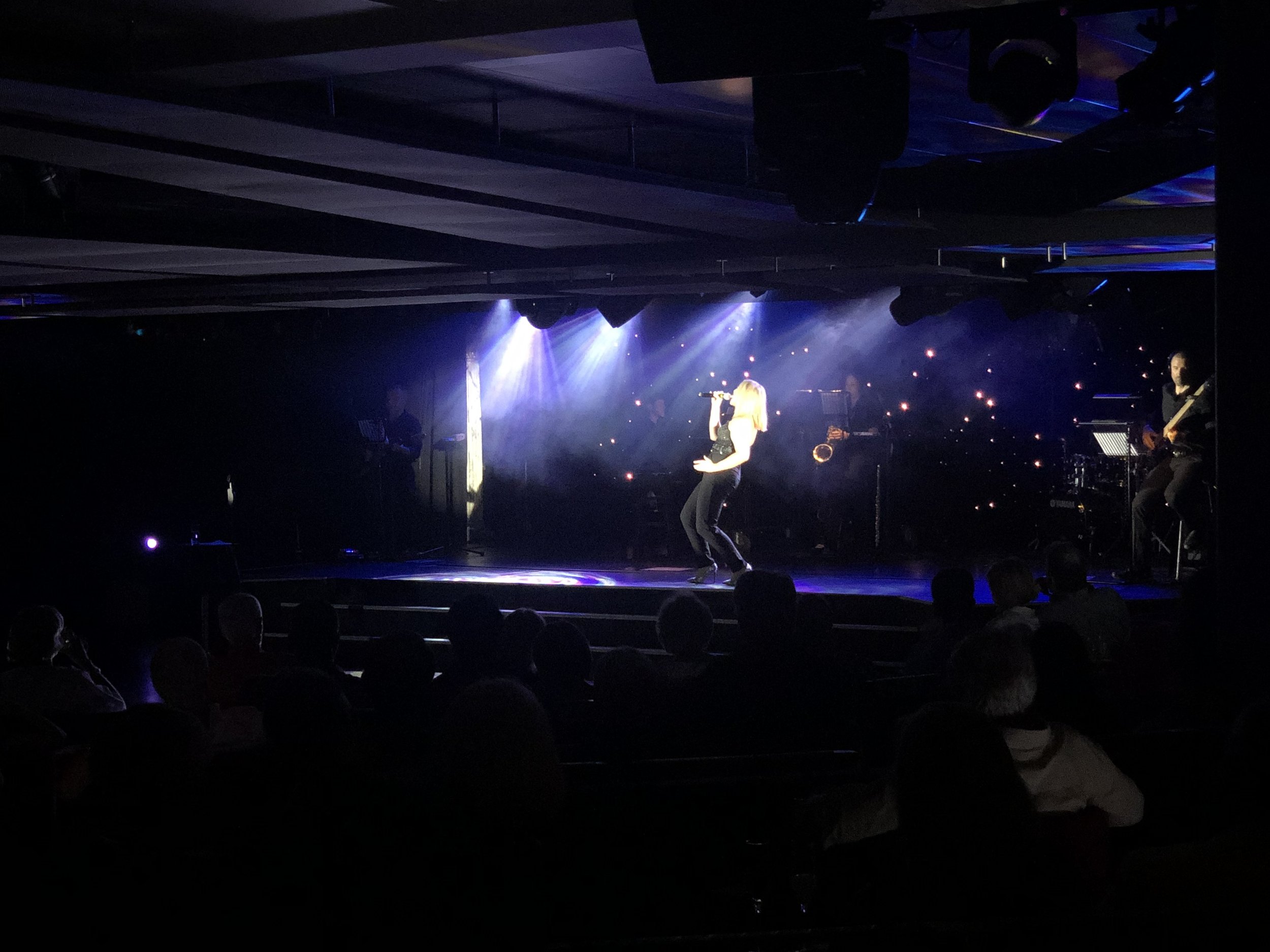 A woman singing on a stage with microphones and musical instruments, illuminated by stage lights, with an audience in the foreground.
