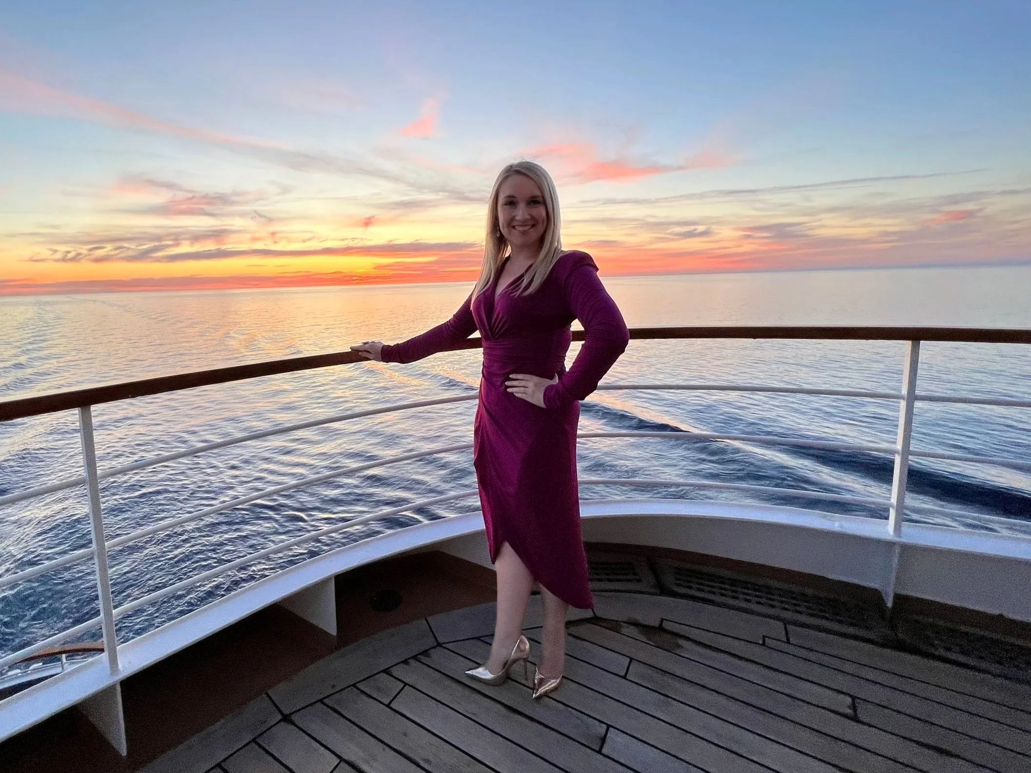 A woman in a purple dress and gold heels standing on a boat deck, smiling with her hand on her hip, during sunset over the water.