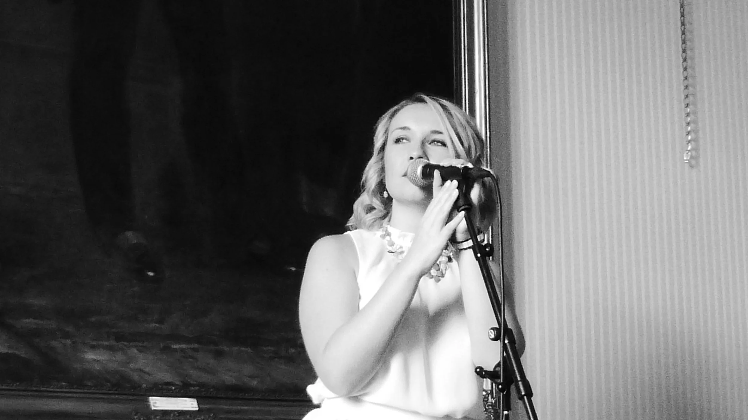 A young woman with wavy hair singing into a microphone, standing against a wall with vertical stripes in a black-and-white photo.