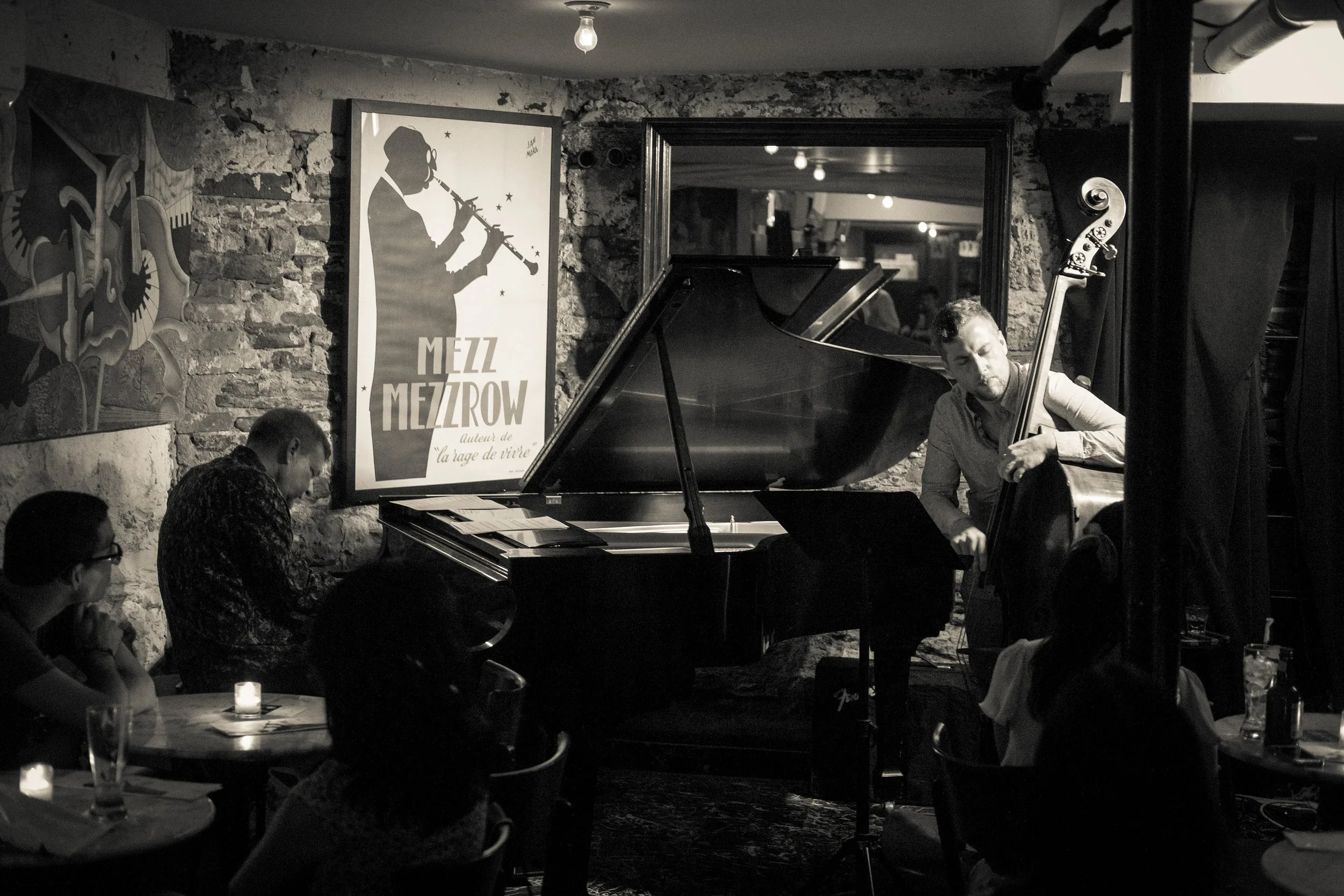 A black and white photo of a jazz performance at a dimly lit venue with exposed brick walls. A man is playing the piano while another man is playing the double bass. There are some audience members sitting at tables, and a poster on the wall reads 'M