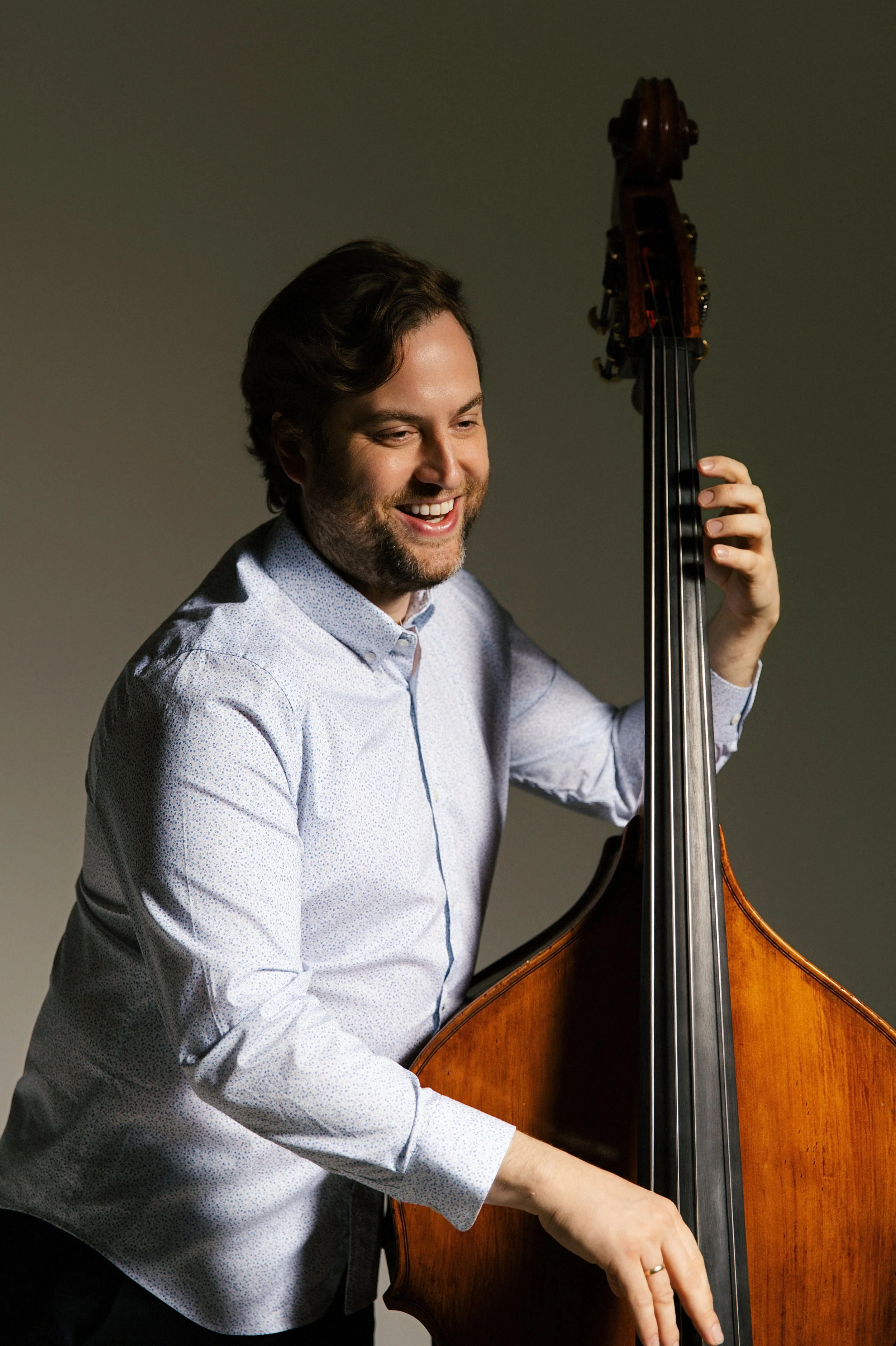 A man with dark hair and a beard smiling while playing a double bass. He is wearing a white patterned shirt and is positioned against a plain gray background.