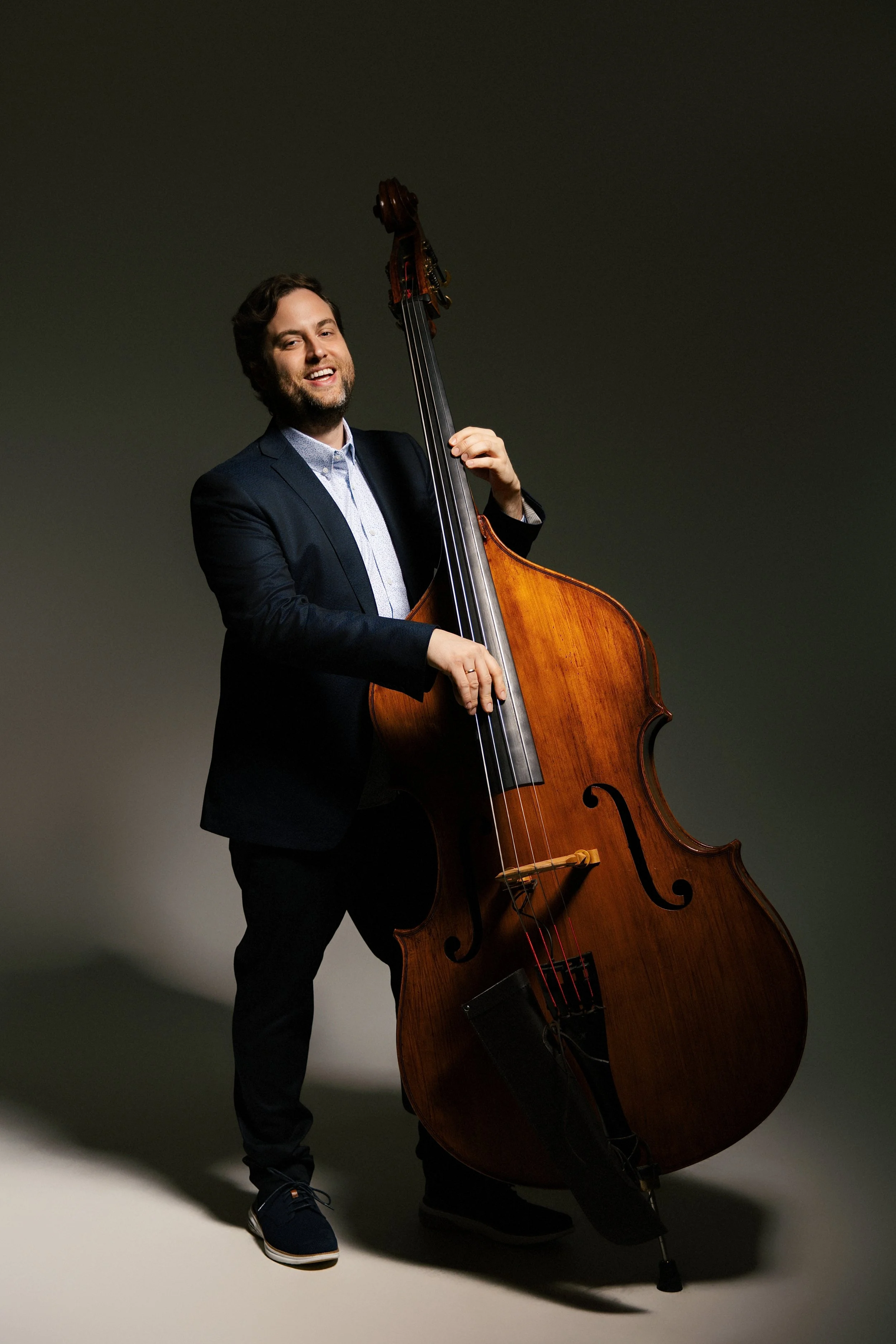 A man in a dark suit playing a large wooden double bass, standing against a dark background.