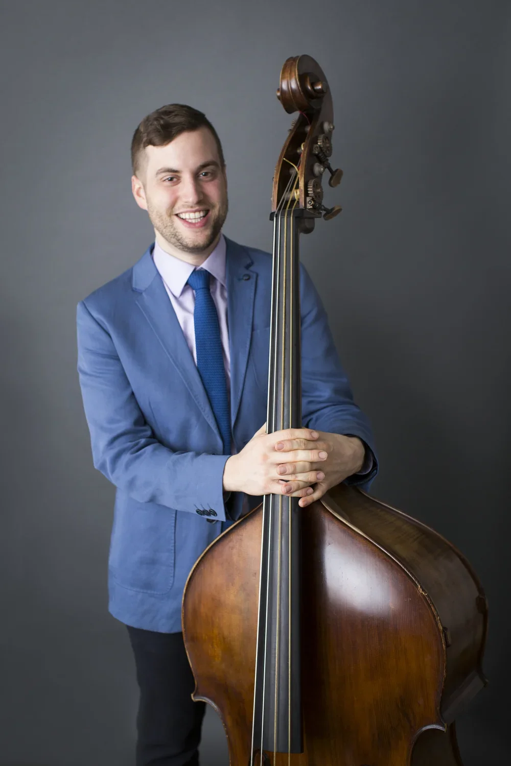 A young man in a blue suit holding a double bass, smiling at the camera, against a plain gray background.