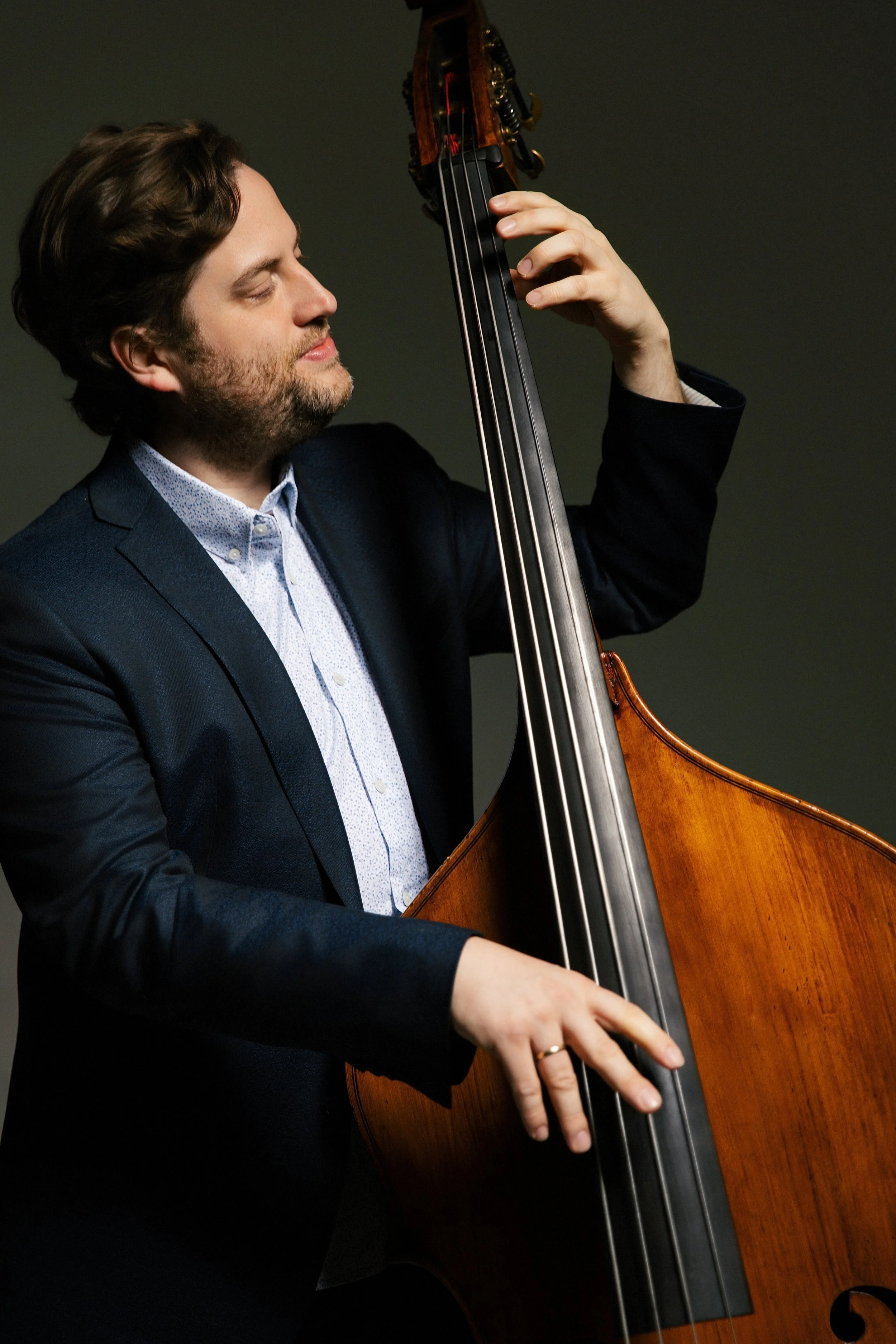 A man with brown hair and a beard, wearing a dark suit with a light patterned shirt, is playing a double bass with a focused expression, standing against a dark background.