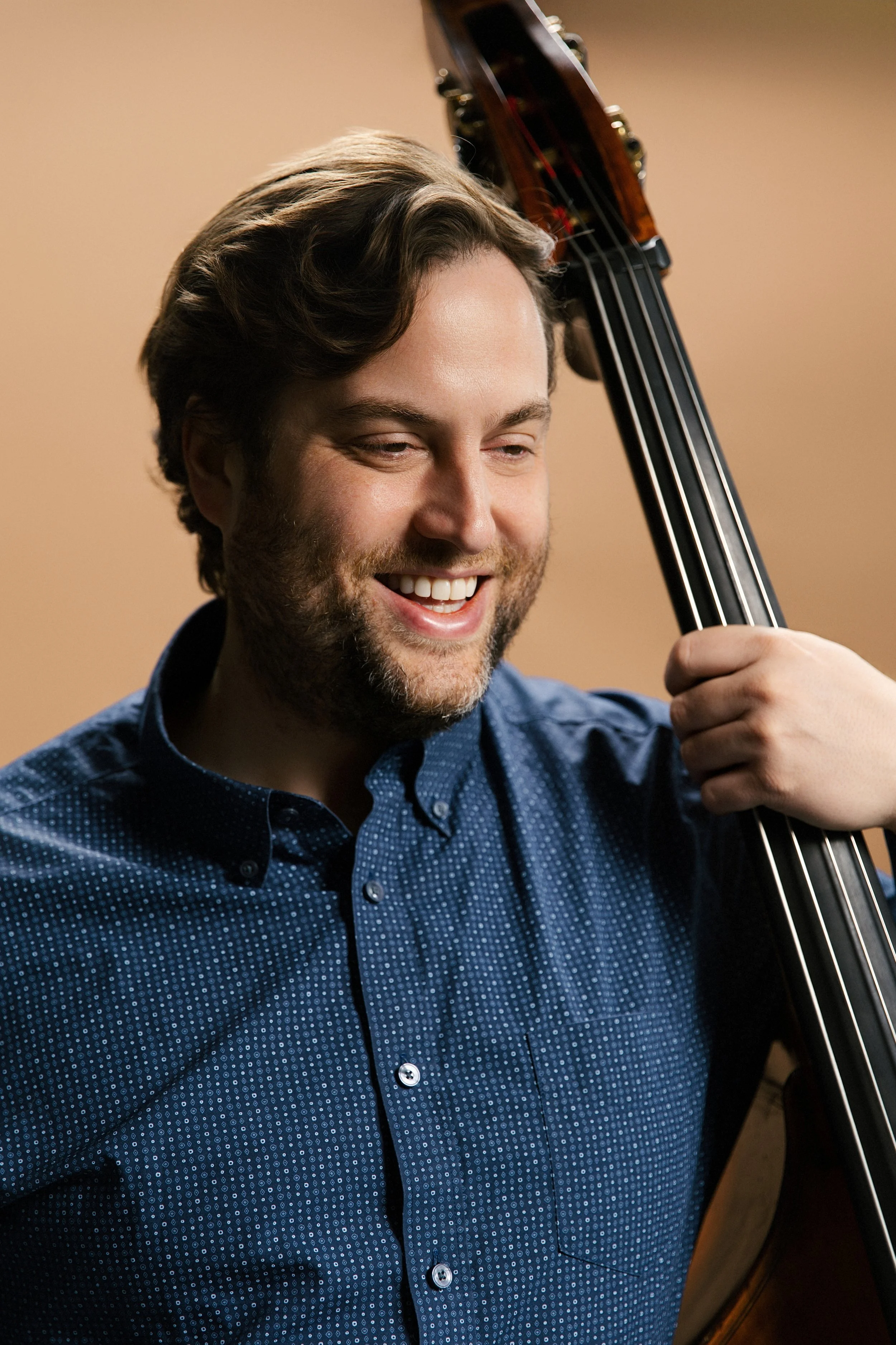 A man with brown hair and a beard, smiling, holds a double bass next to his face.