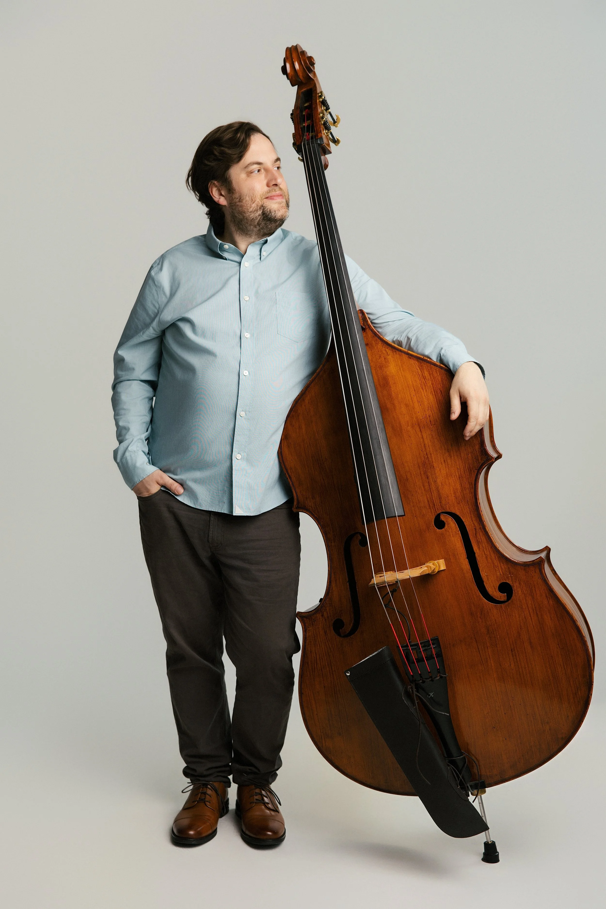 A man with brown hair and a beard wearing a light blue button-up shirt, dark pants, and brown shoes, standing against a plain light gray background, holding a large wooden double bass.