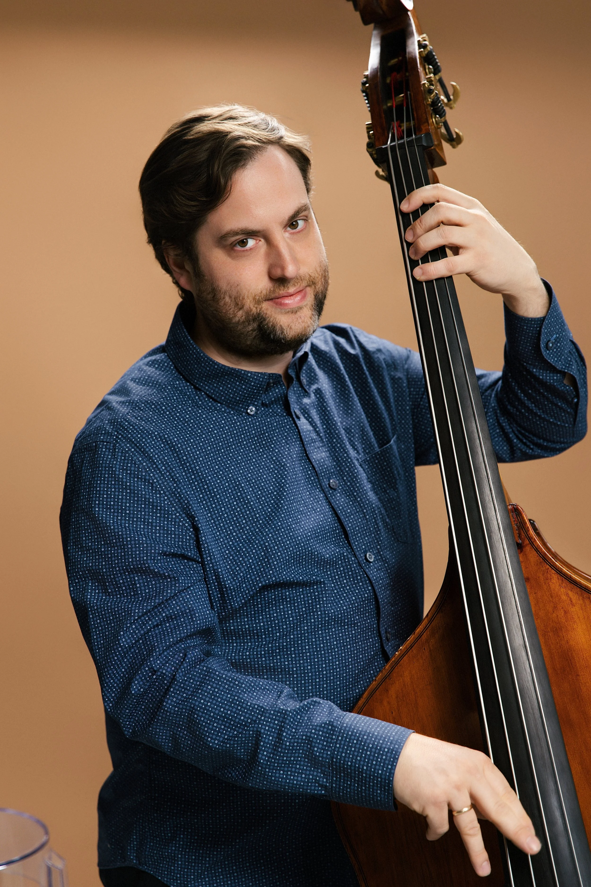 A man with brown hair and a beard wearing a blue patterned shirt is playing a double bass.