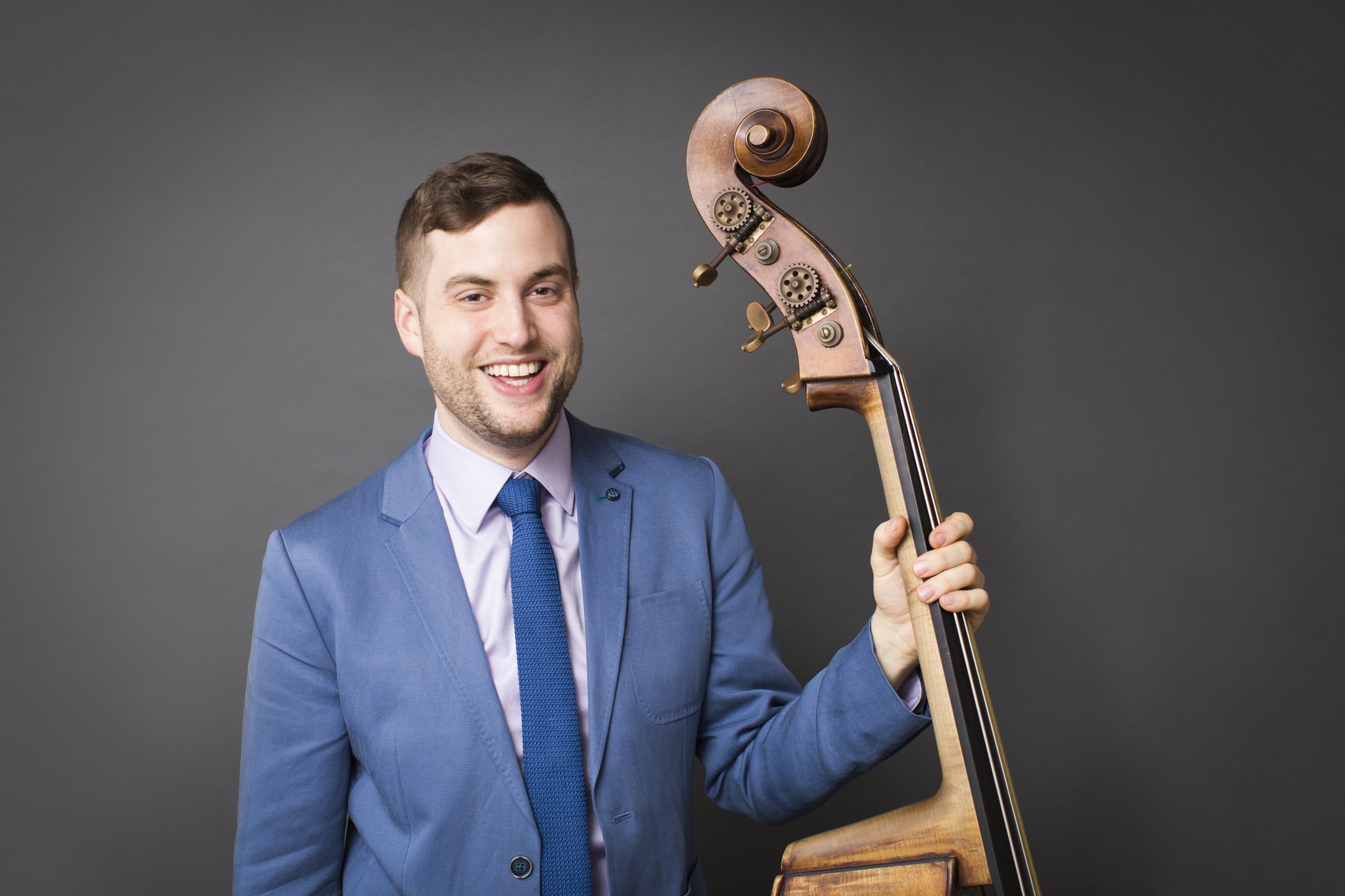 A smiling man in a blue suit holding a violin in front of a gray background.