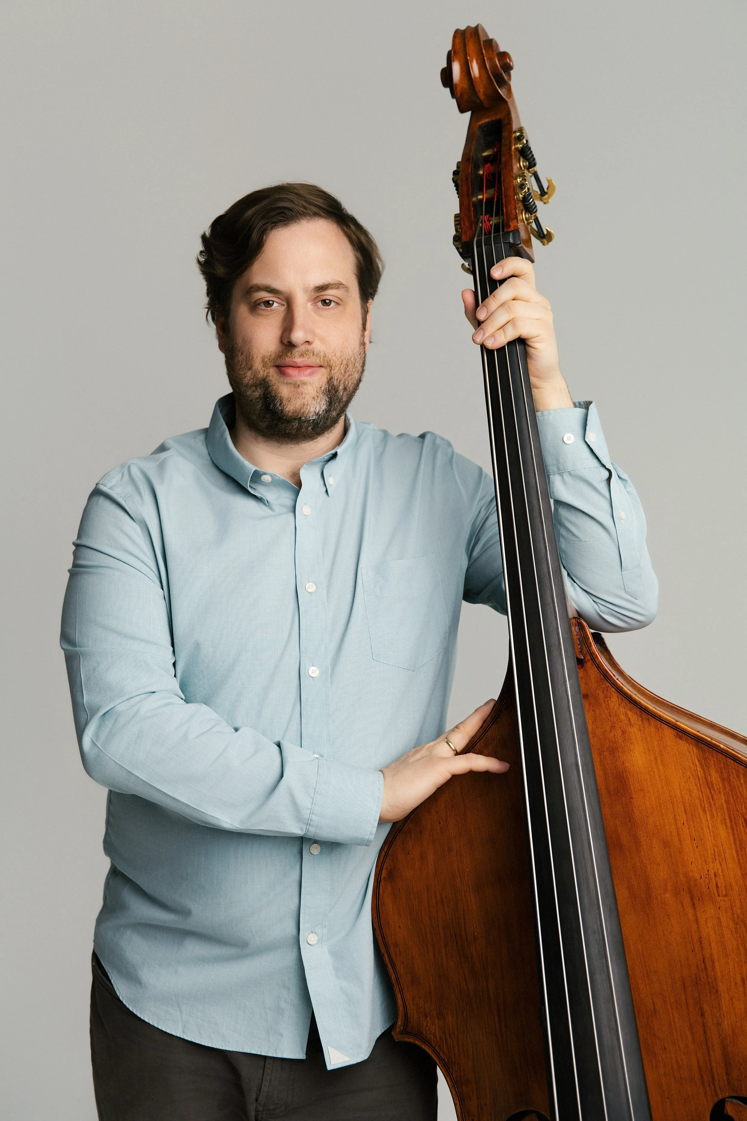 A man with dark hair and beard wearing a light blue button-up shirt, standing against a plain background, holding a large wooden upright bass.