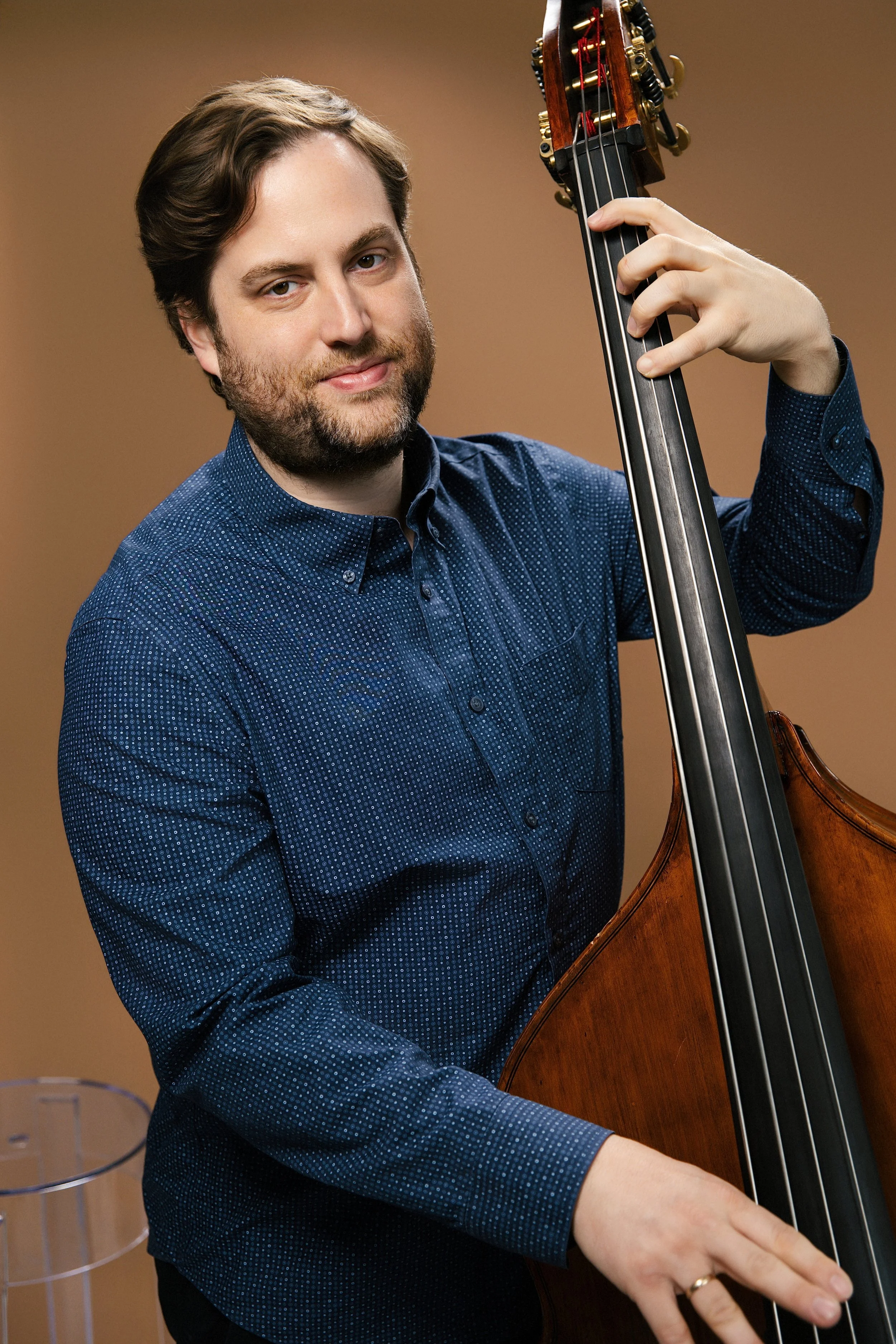 A man with brown hair and facial hair playing a double bass, wearing a blue patterned shirt, against a plain brown background.