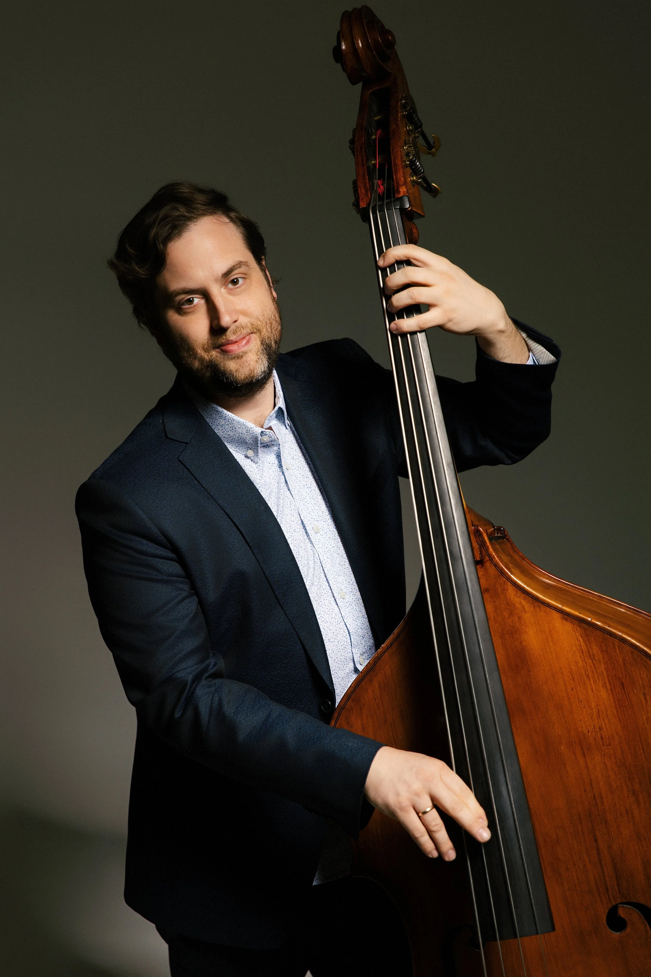 A man wearing a dark suit and light blue shirt playing an upright bass against a plain gray background.