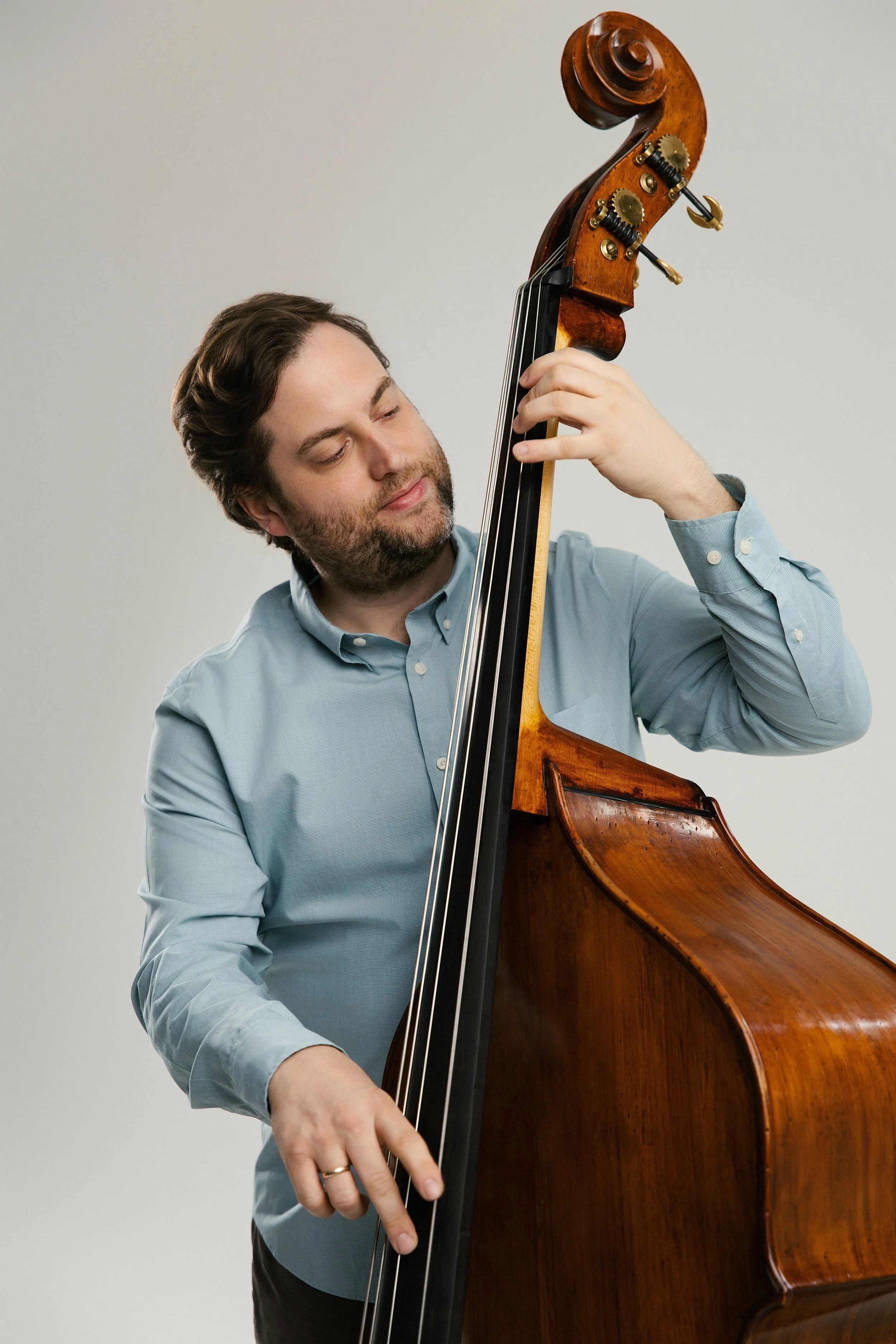 A man with dark hair and a beard playing a double bass. He is wearing a light blue shirt and appears focused on playing the instrument.
