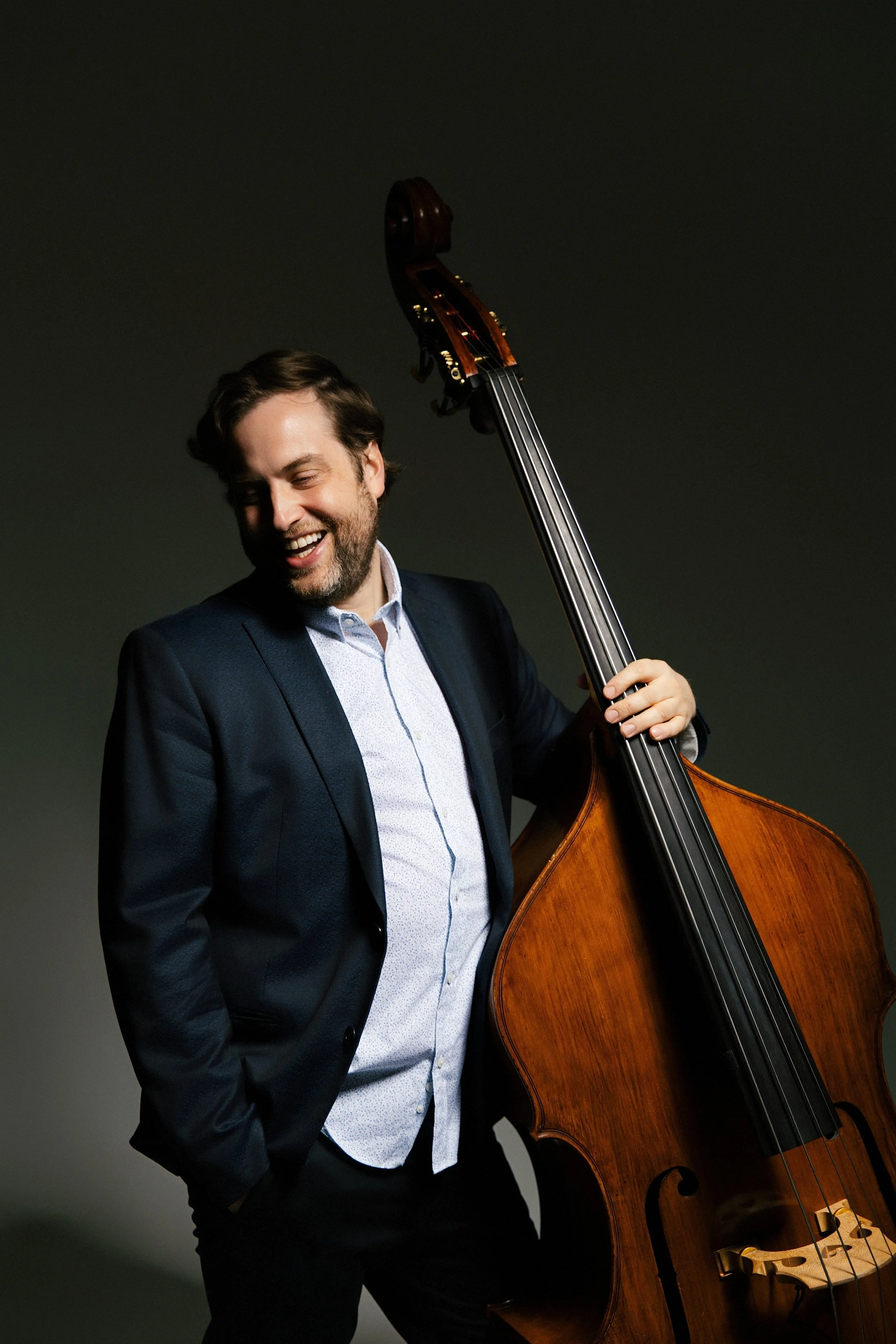 A man with dark hair and a beard, dressed in a blue suit and light blue shirt, holding a wooden double bass, smiling against a dark background.