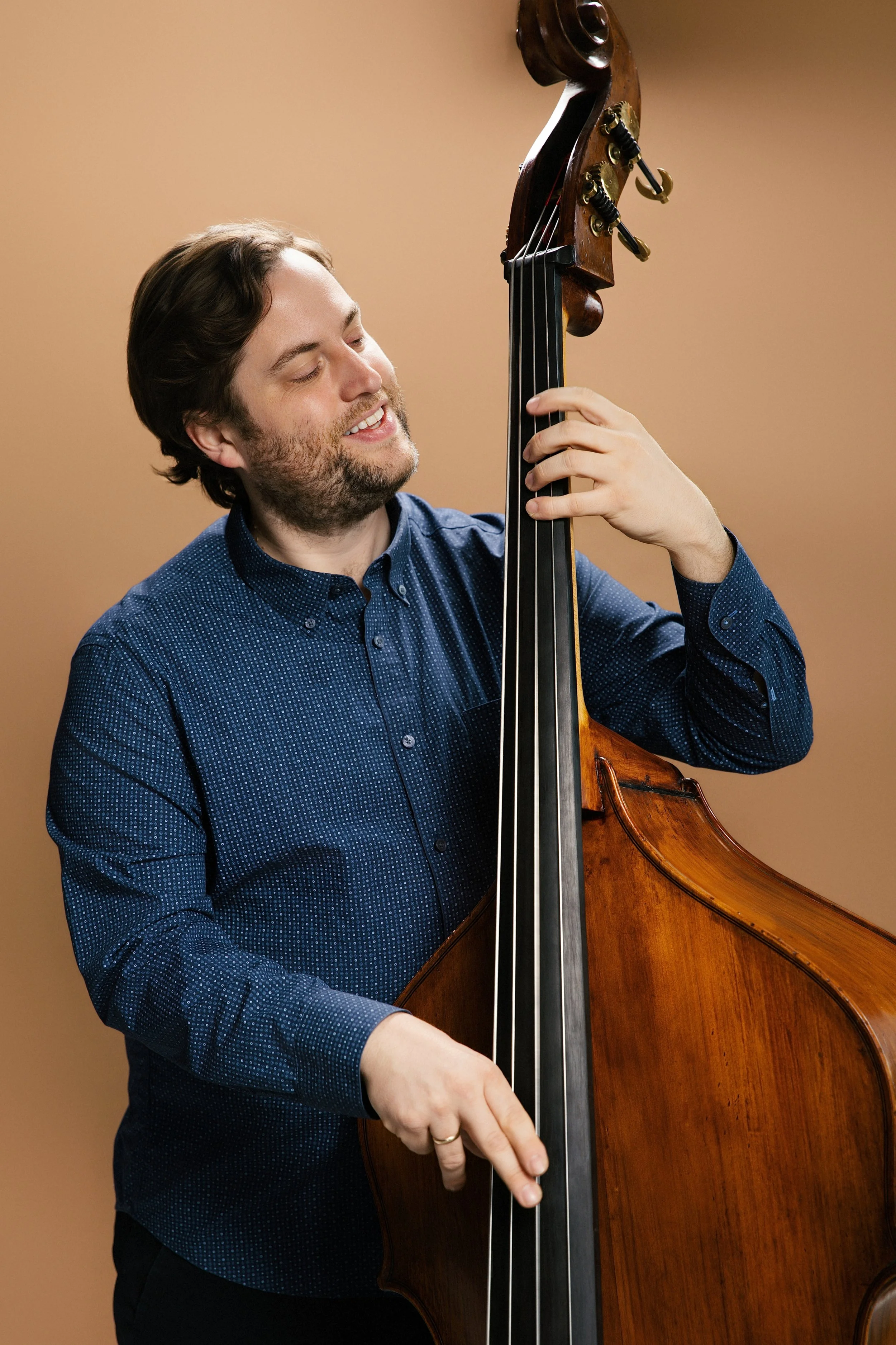A man with brown hair and a beard playing a double bass indoors against a plain brown background.