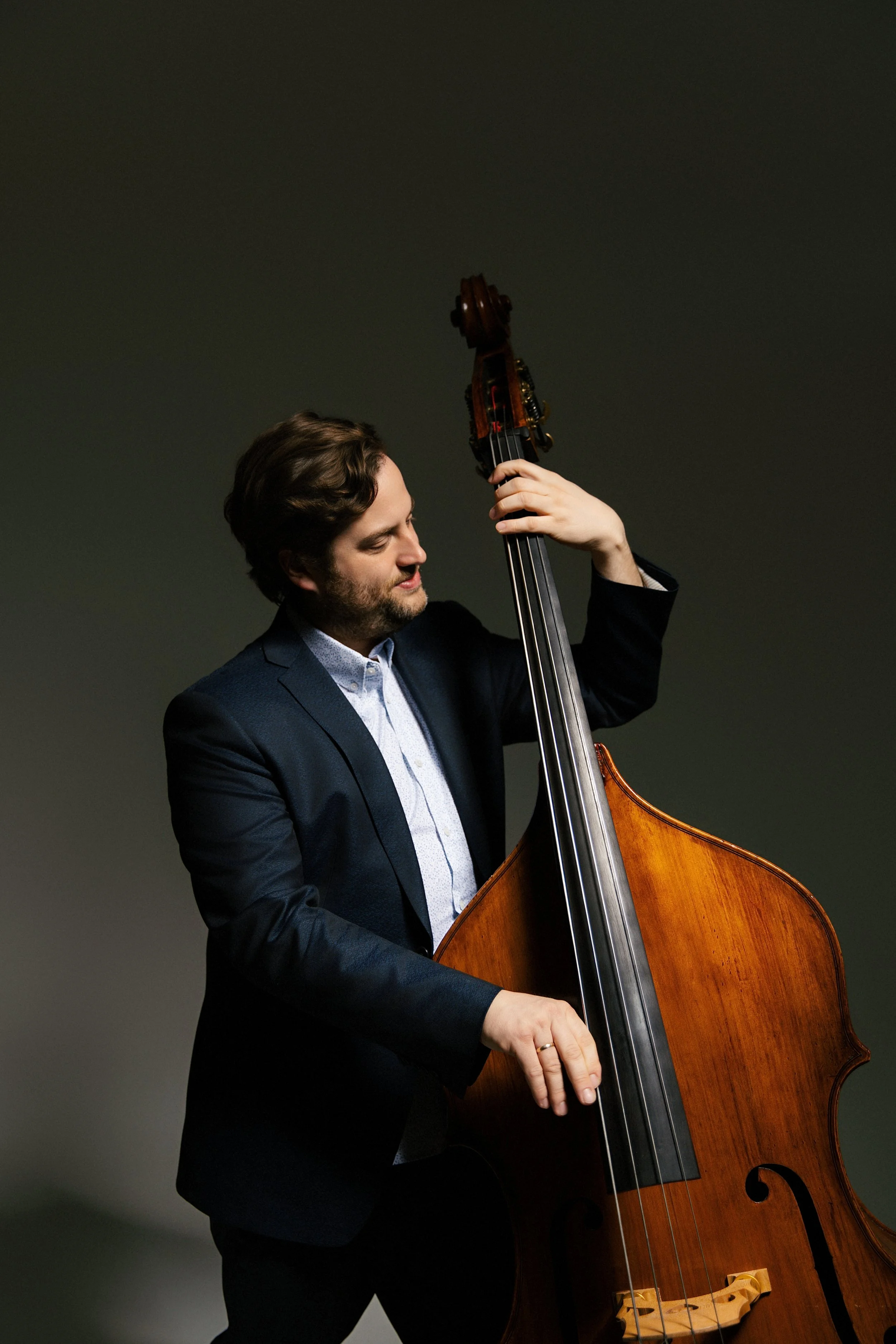 A man with brown hair and a beard playing a double bass against a dark background.