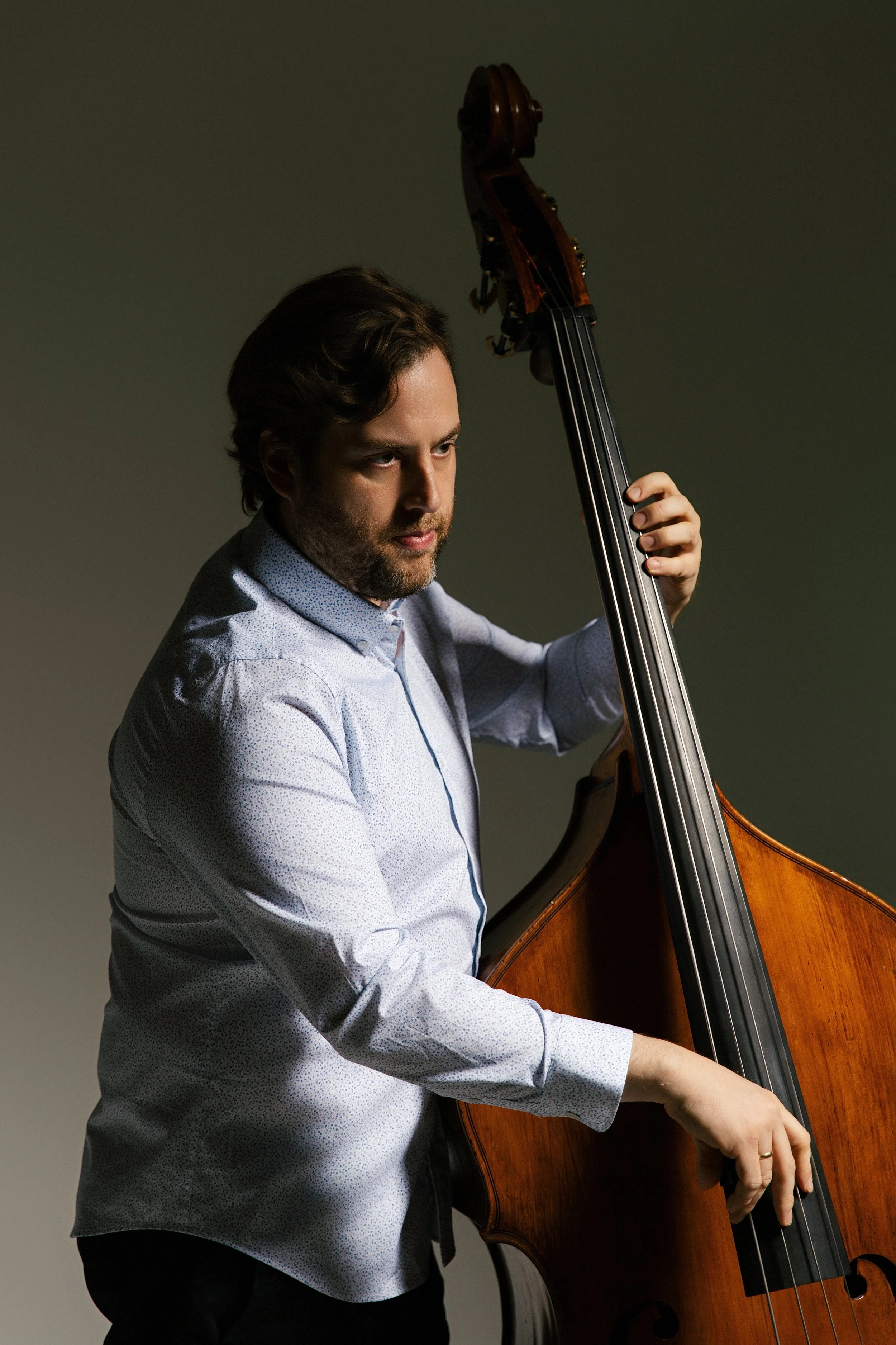 A man with dark hair and a beard playing a double bass against a plain background.