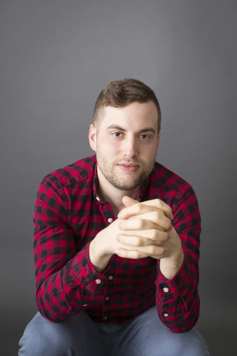 Portrait of a young man with short brown hair and a beard, wearing a red and black checkered shirt, sitting against a plain gray background, with his hands clasped together and looking into the camera.
