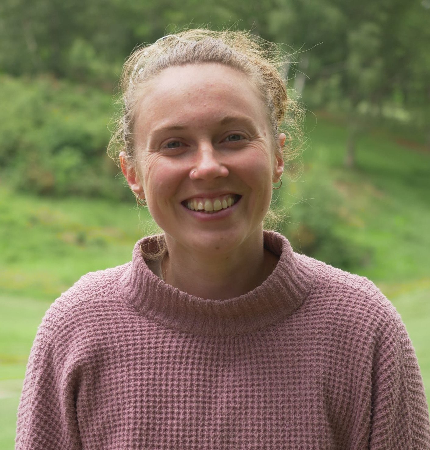 Lucy Gray - A smiling woman with blonde hair tied back, wearing a pink textured sweater, standing outdoors with a blurred green landscape in the background.