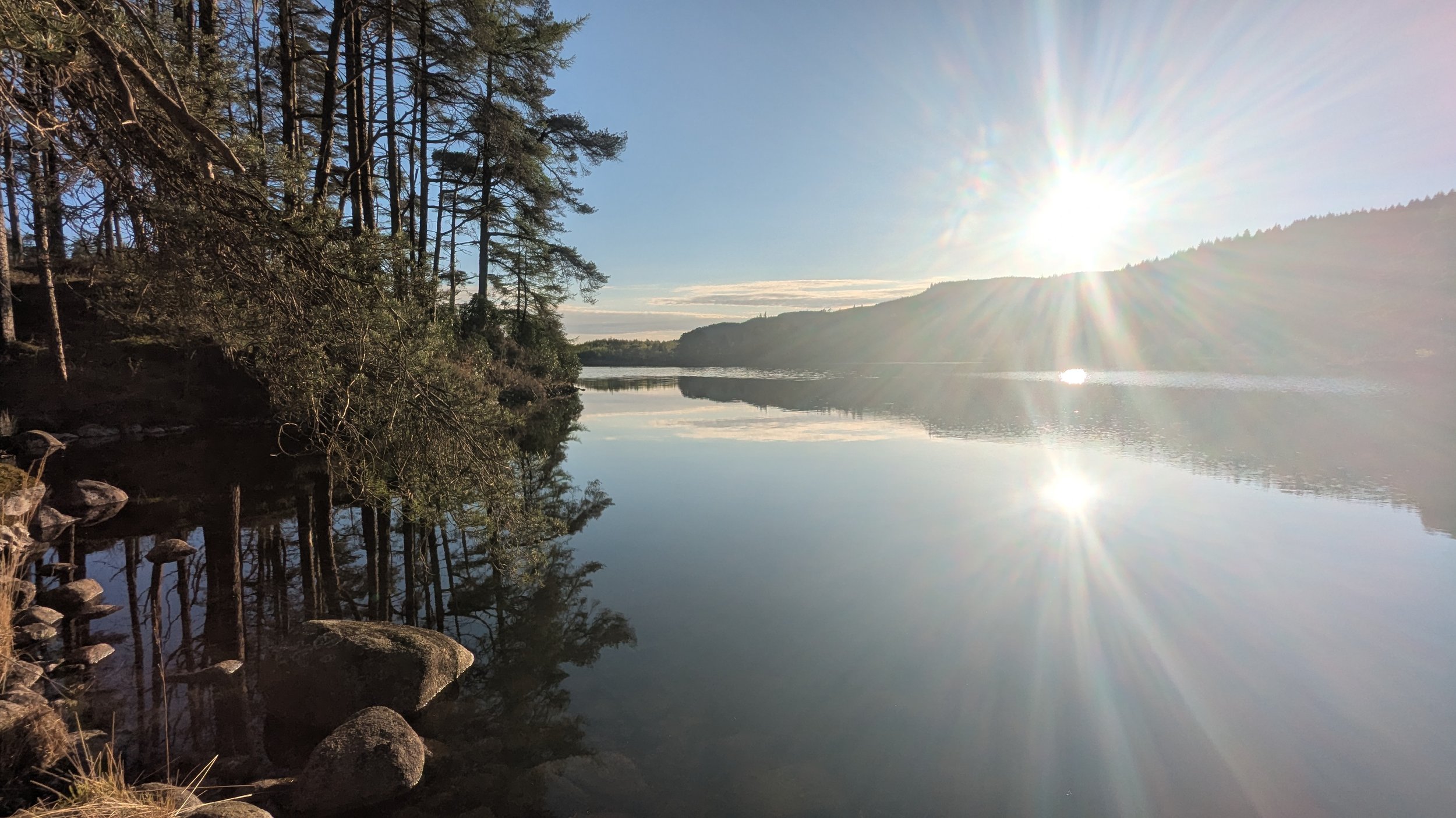 A serene lakeside scene with calm water reflecting the sky, surrounded by trees on the left and hills on the right, with the sun shining brightly from the sky.