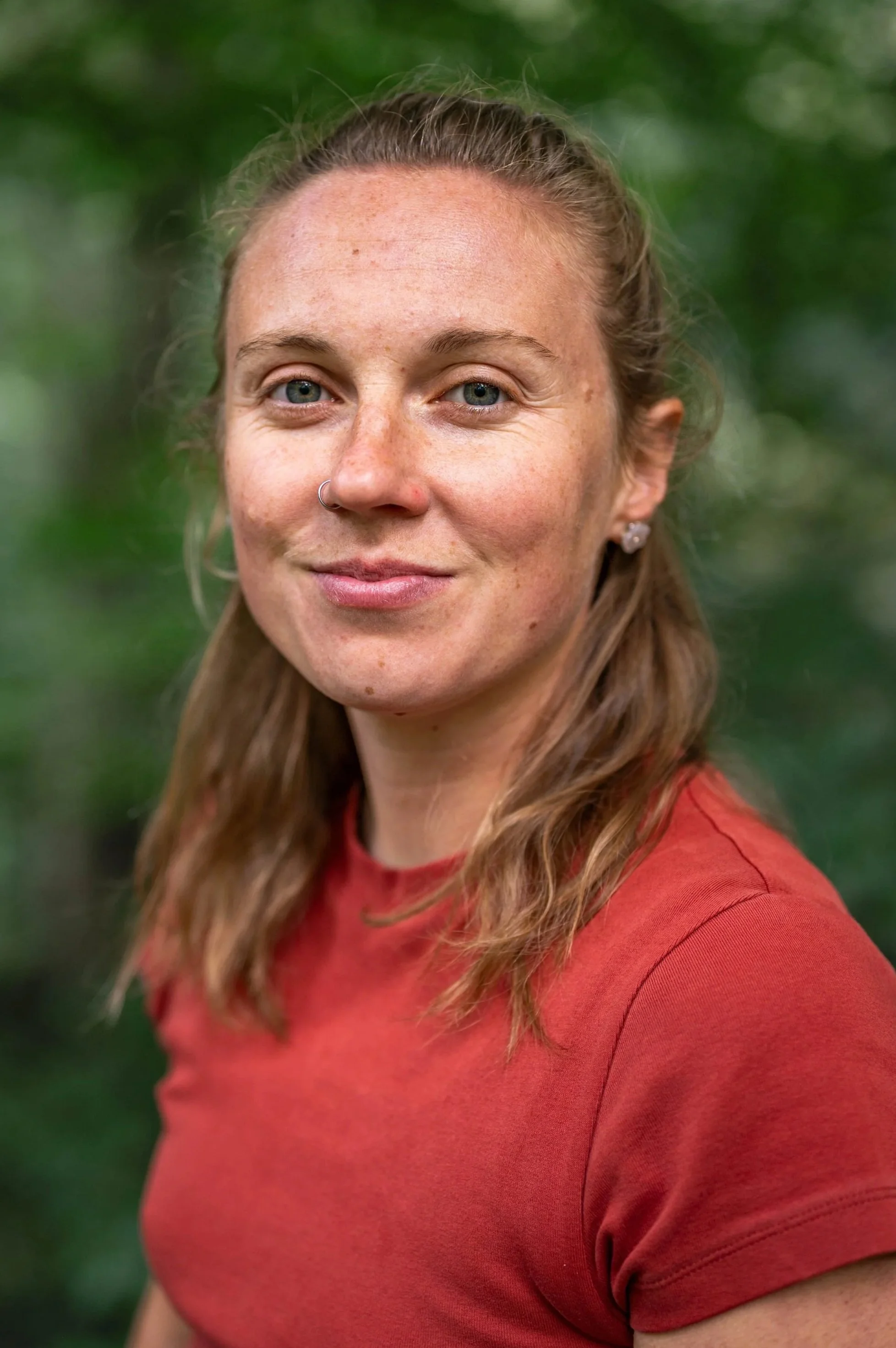 Close-up of a woman with blue eyes, red hair, a nose ring, and earrings, wearing a red top against a blurred green outdoor background.