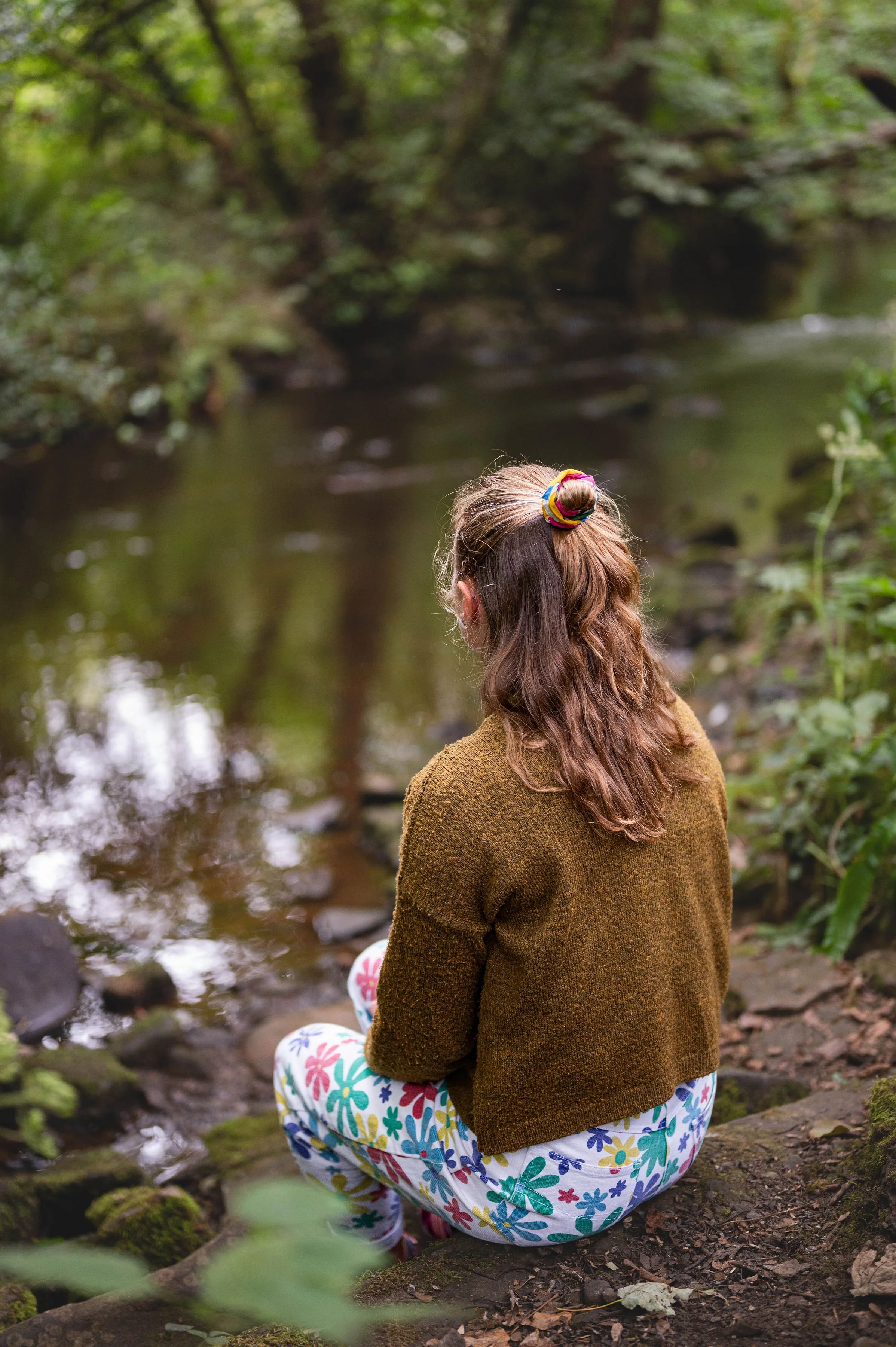 A person with long brown hair, wearing a colorful hair scrunchie, a brown sweater, and brightly patterned pants, kneeling on the ground near a stream in a forest with green foliage.