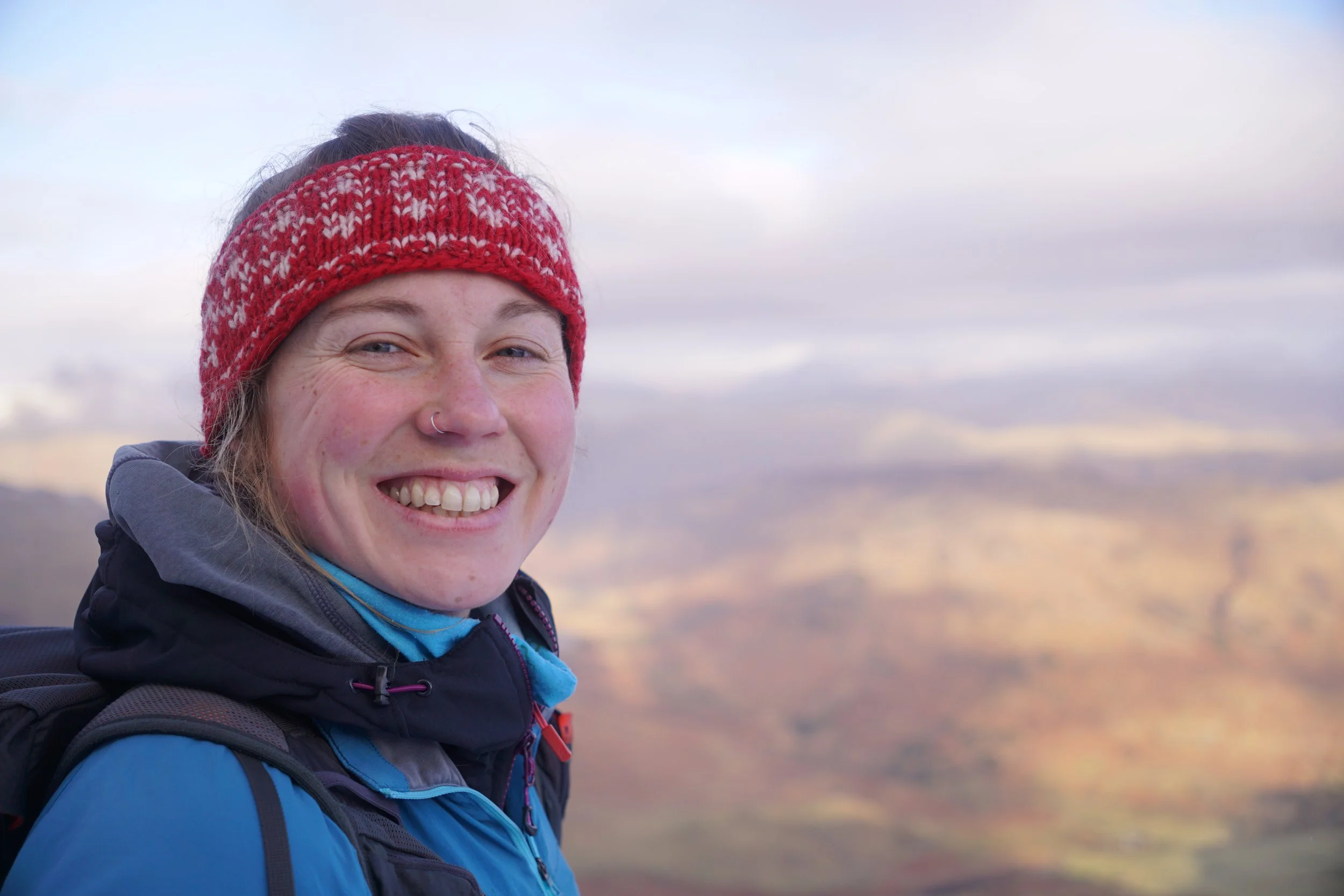 Lucy Gray - A smiling woman wearing a red and white knit headband and blue outdoor jacket, standing outdoors with a cloudy sky in the background.