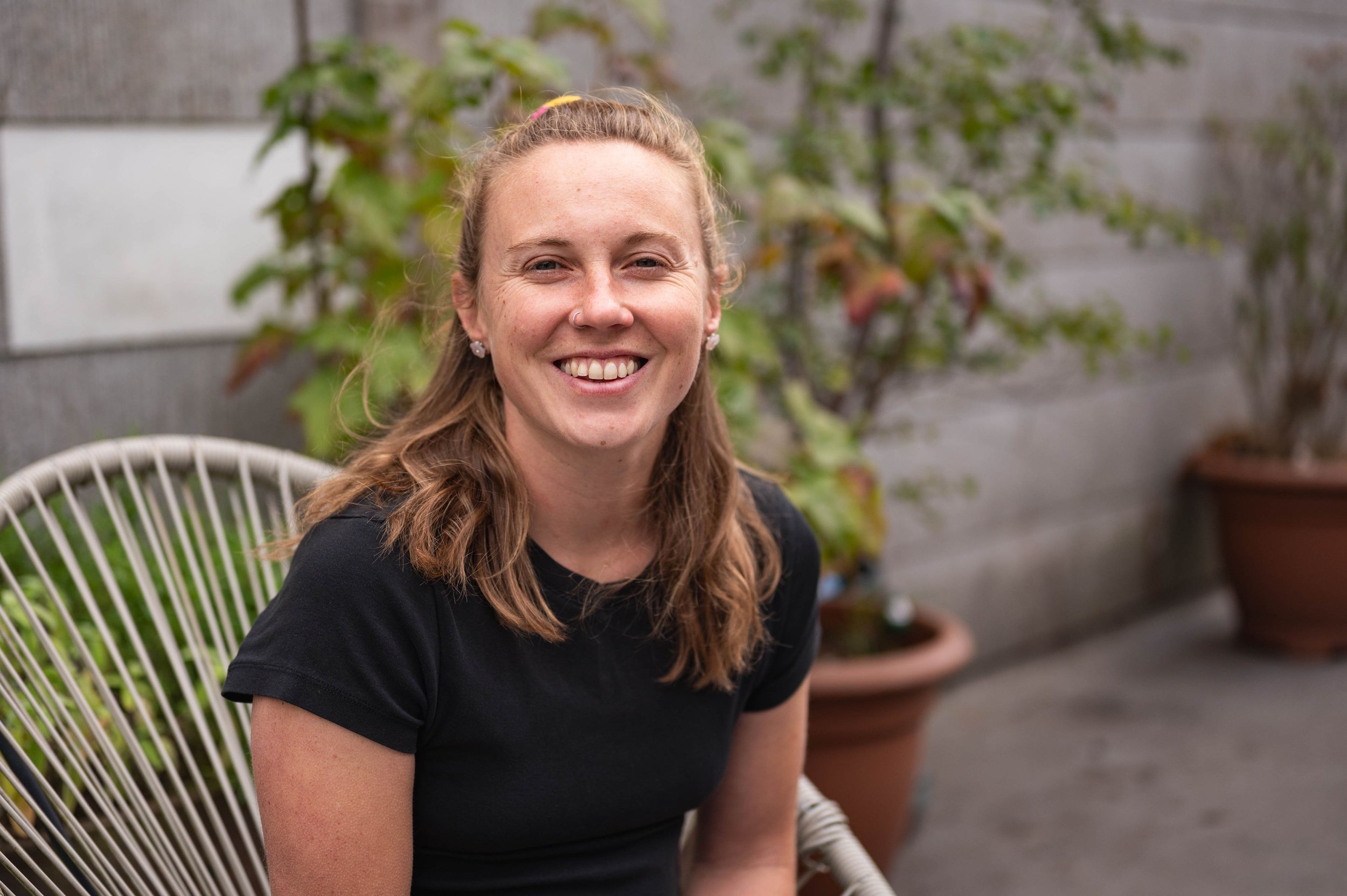 A young woman with long, wavy brown hair and a nose piercing smiling while sitting outdoors on a white wire chair with potted plants and greenery in the background.