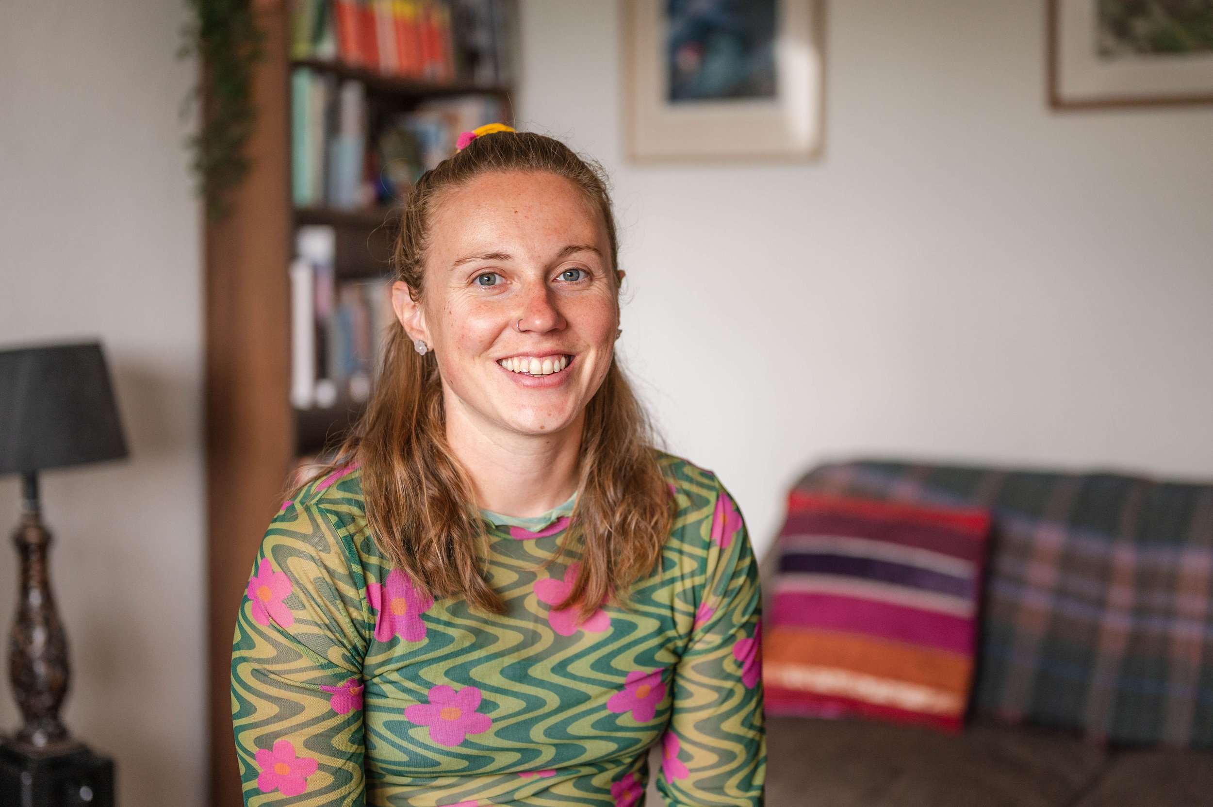 A smiling woman with long reddish hair, blue eyes, and a nose ring, sitting indoors in front of a bookshelf and wall art, wearing a colorful patterned shirt with pink flowers and green swirls.