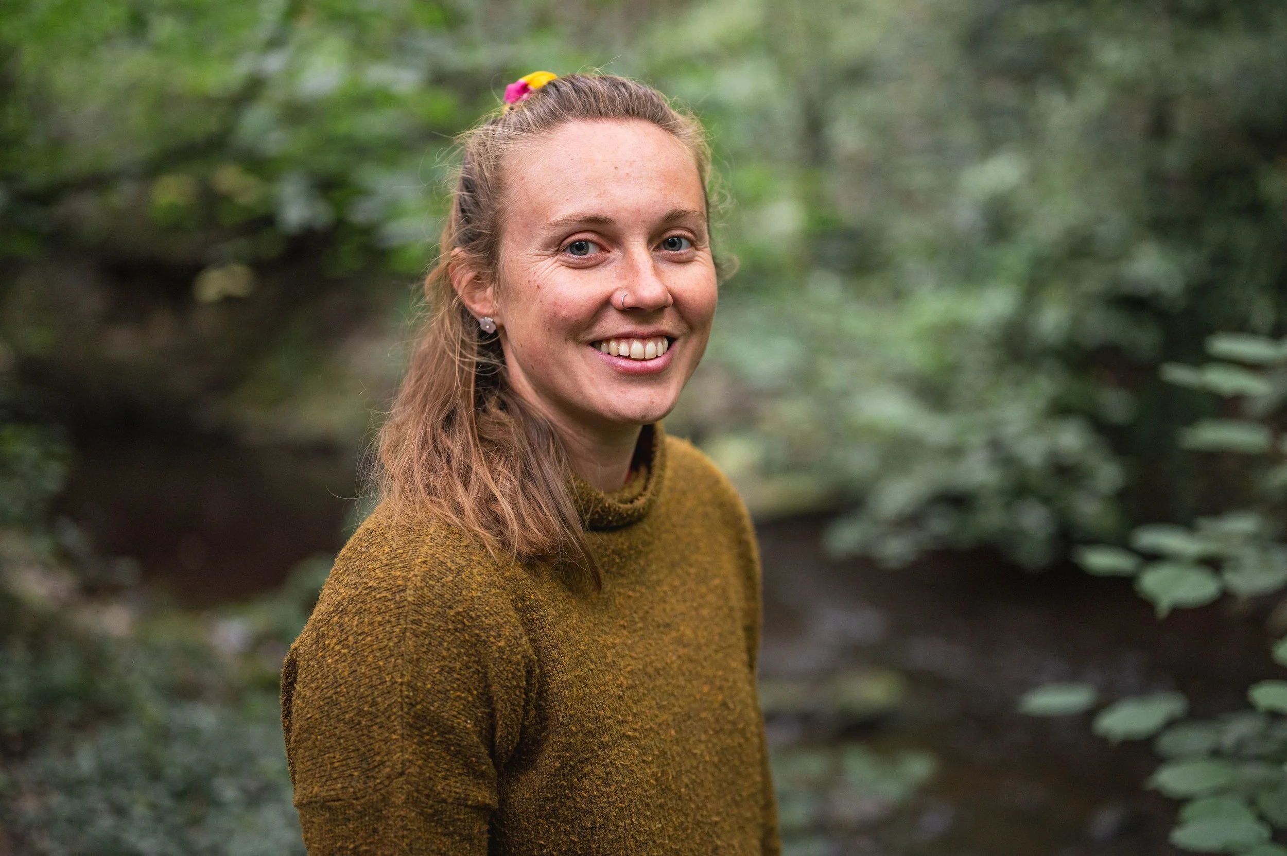 A woman with long hair, wearing a mustard-colored sweater, standing outdoors in a lush green forest, smiling at the camera.